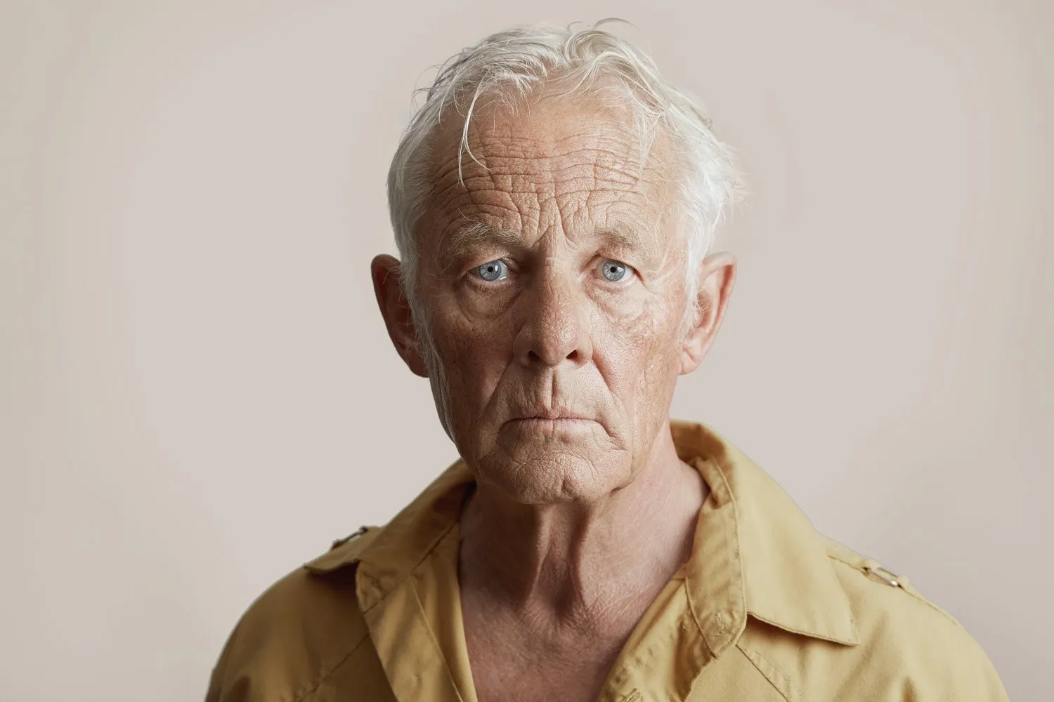 Portrait of an older man with gray hair and blue eyes, wearing a beige shirt, neutral background.
