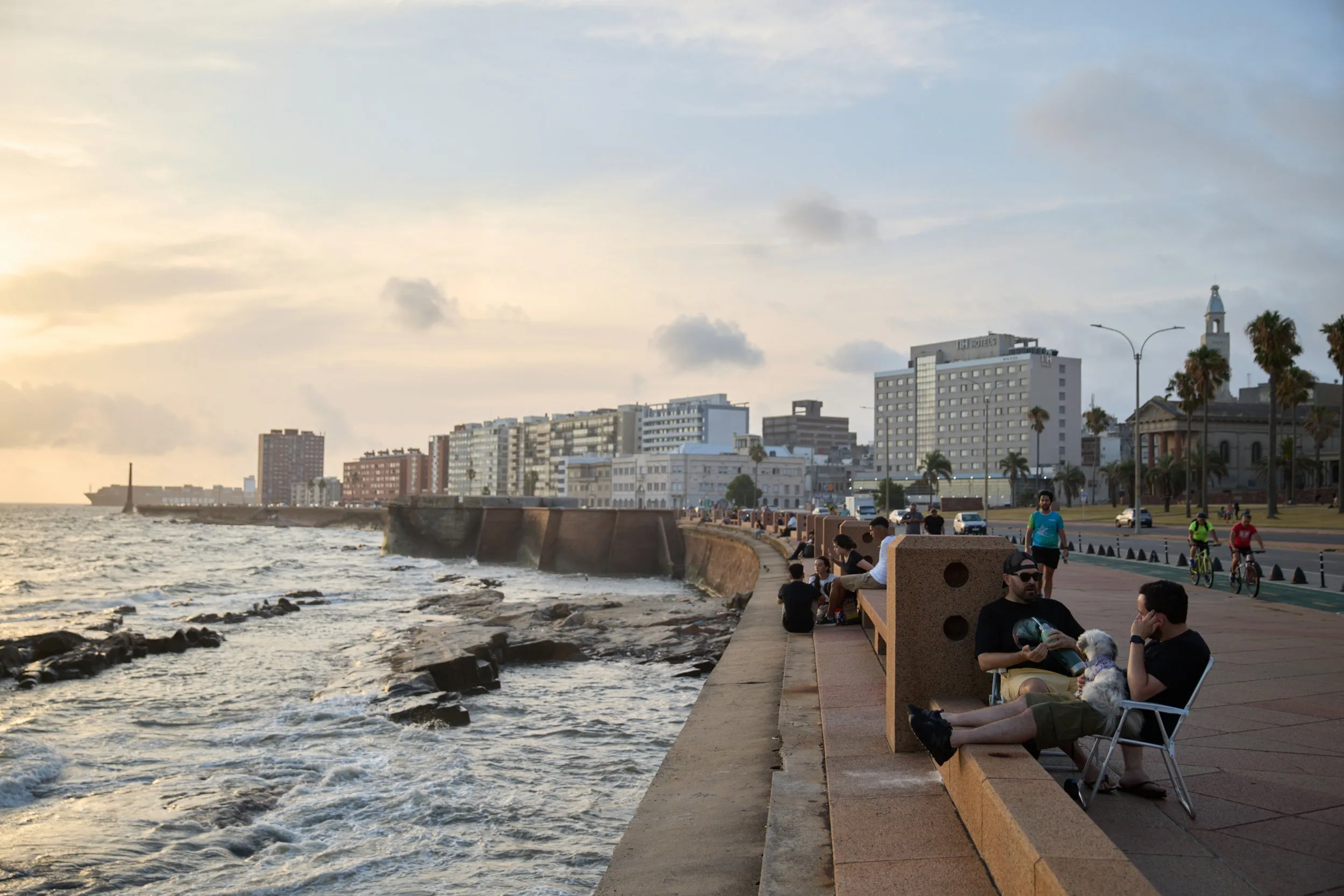 Waves breaking against the rocks of the rambla, people sitting on the benches