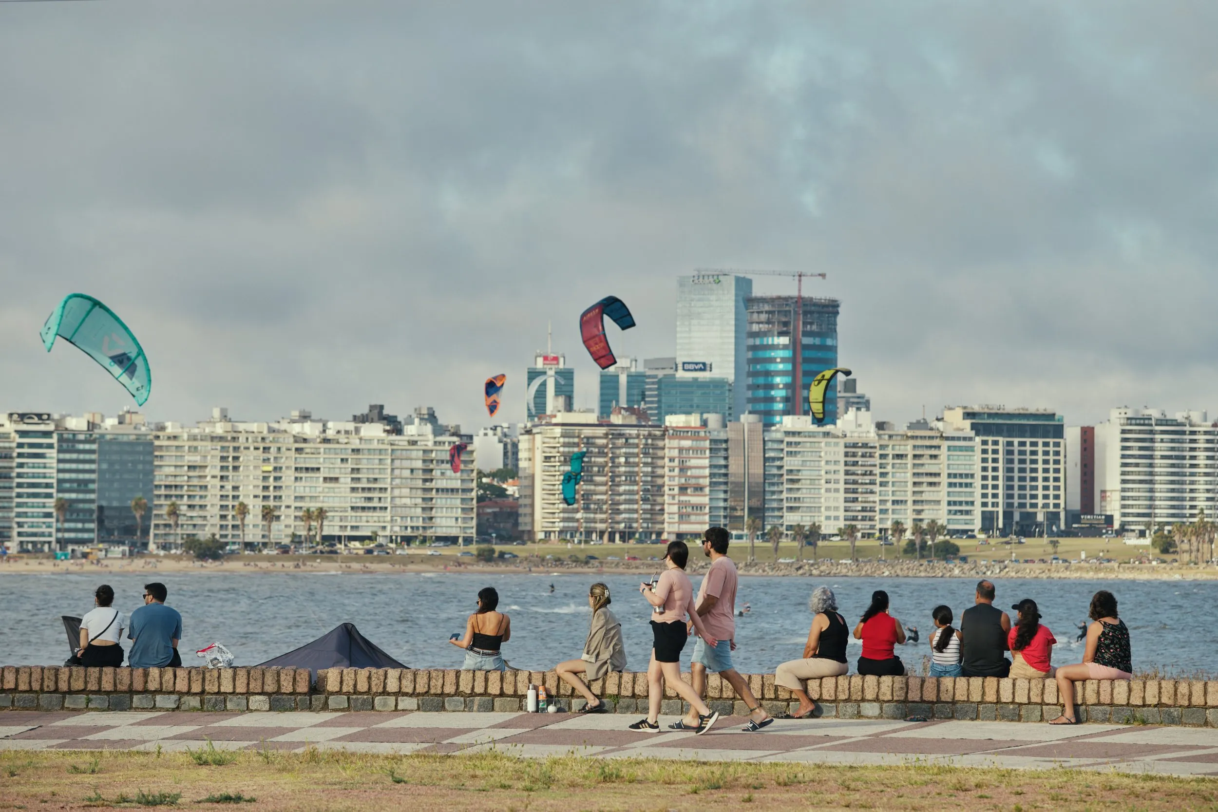 People sitting on the wall of the Rambla de Pocitos, kites over the water and buildings behind