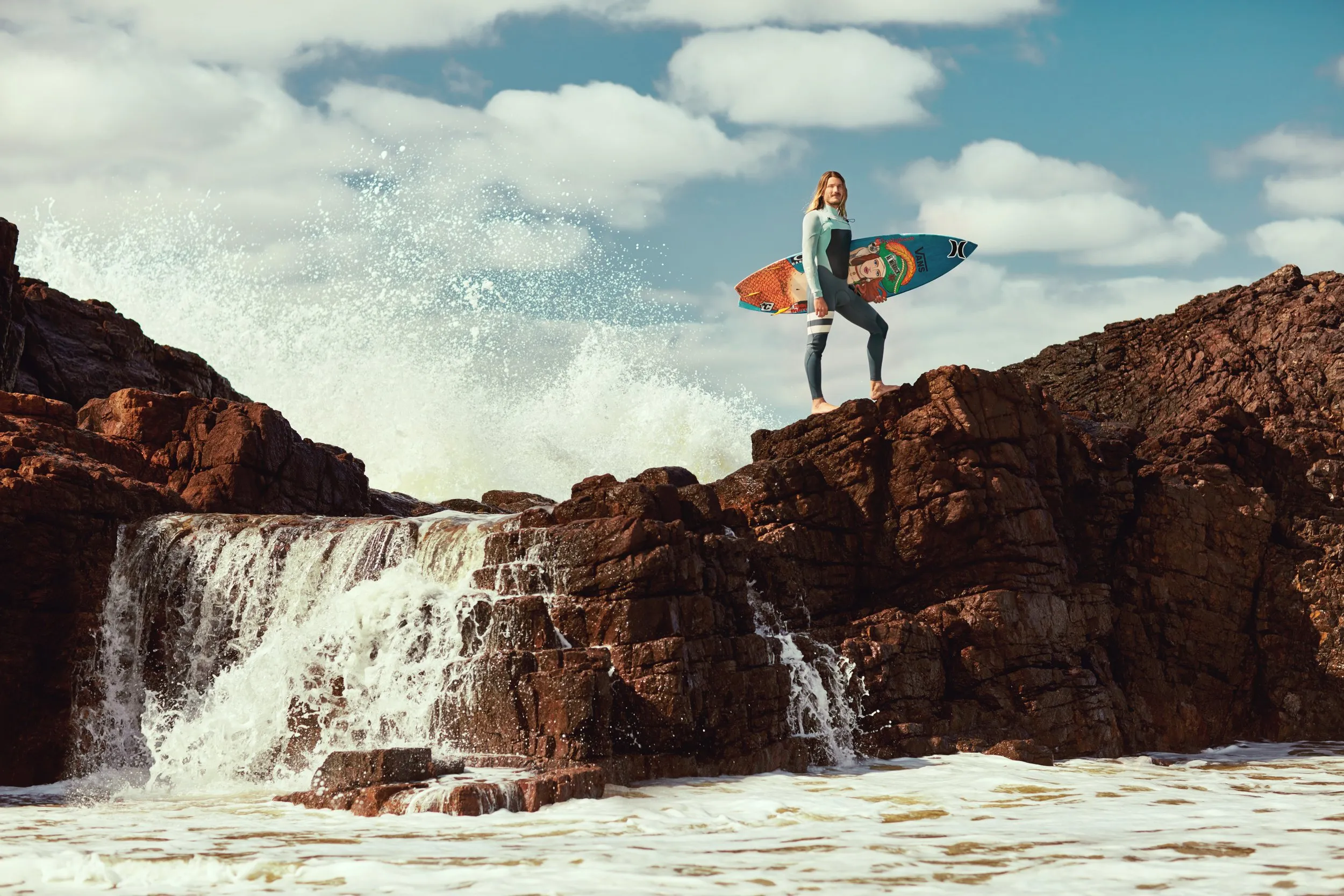 A person in a wetsuit on rocks by the sea, holding a surfboard under their arm, with waves breaking in the background and a partly cloudy sky.