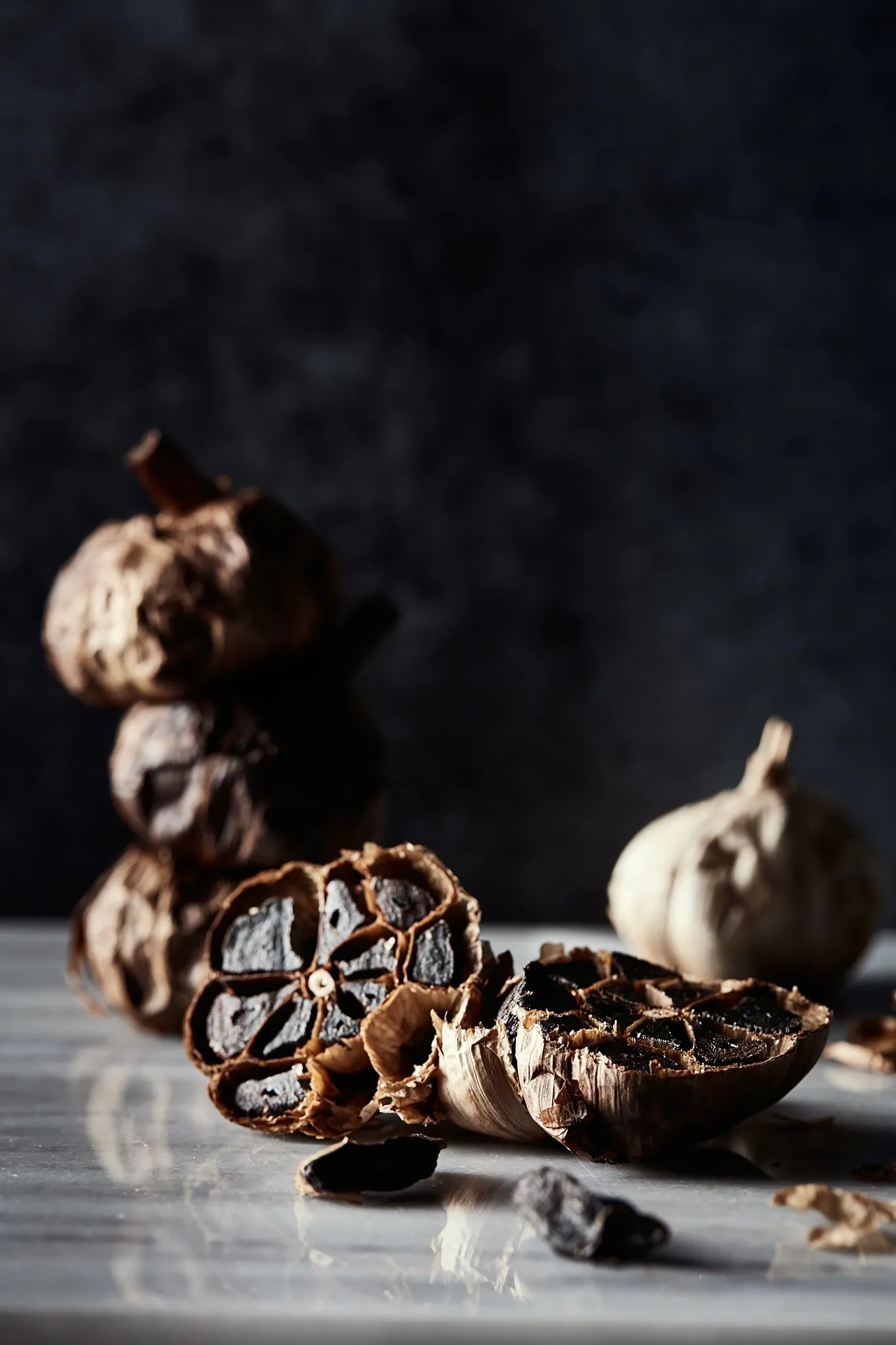Black garlic, dried and in its natural form, with a few fragments on a marble surface against a dark background.