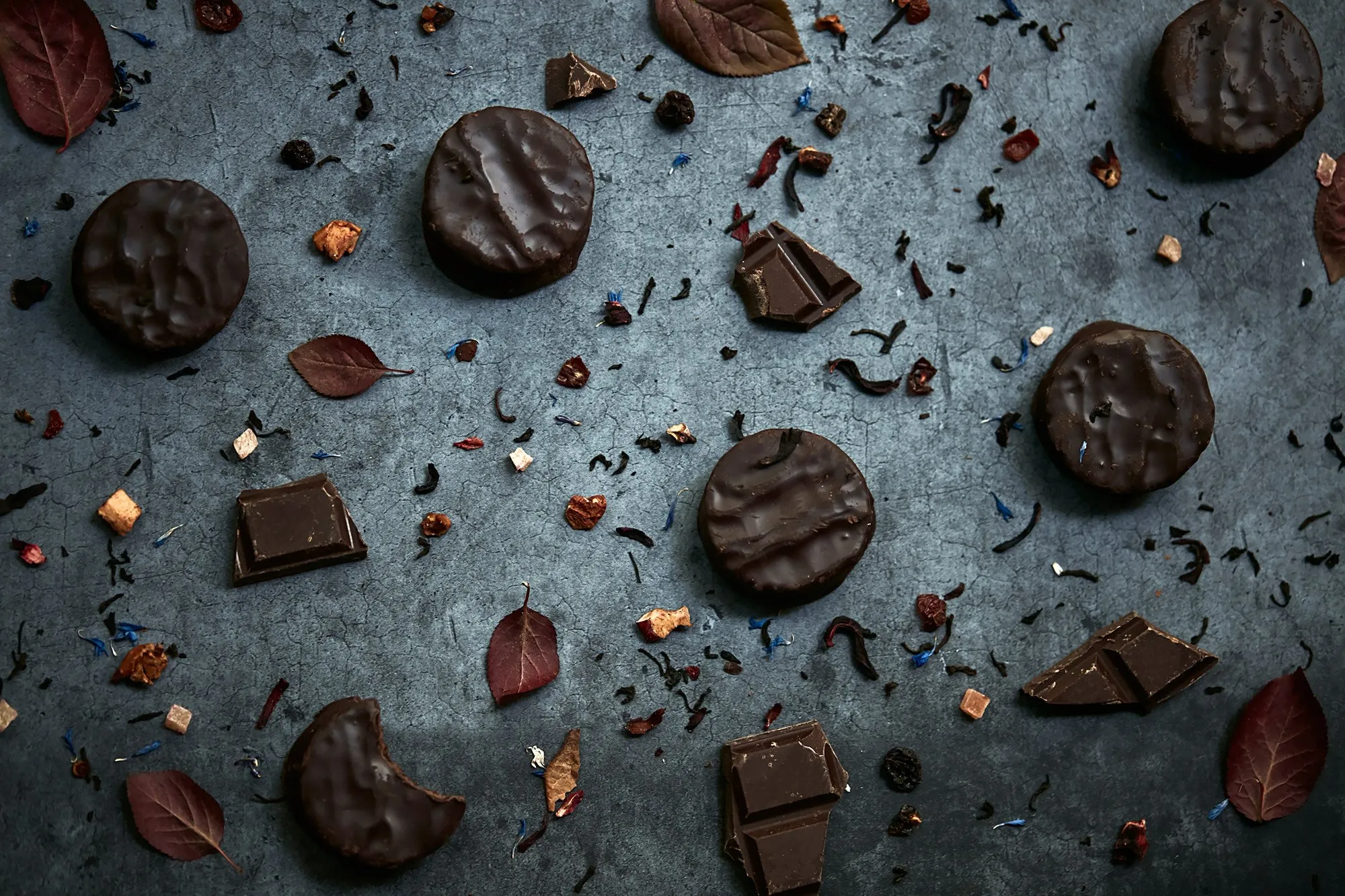 Several wrapped chocolates, some rectangular and some round, with a few dried leaves and small pieces of decorative ingredients scattered on a dark background.