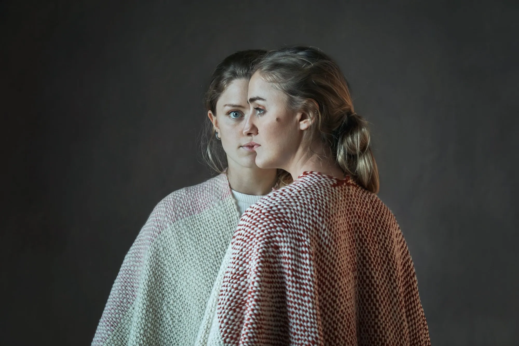 Two women with straight blonde hair, facing each other, wearing knit sweaters, against a dark gray background.