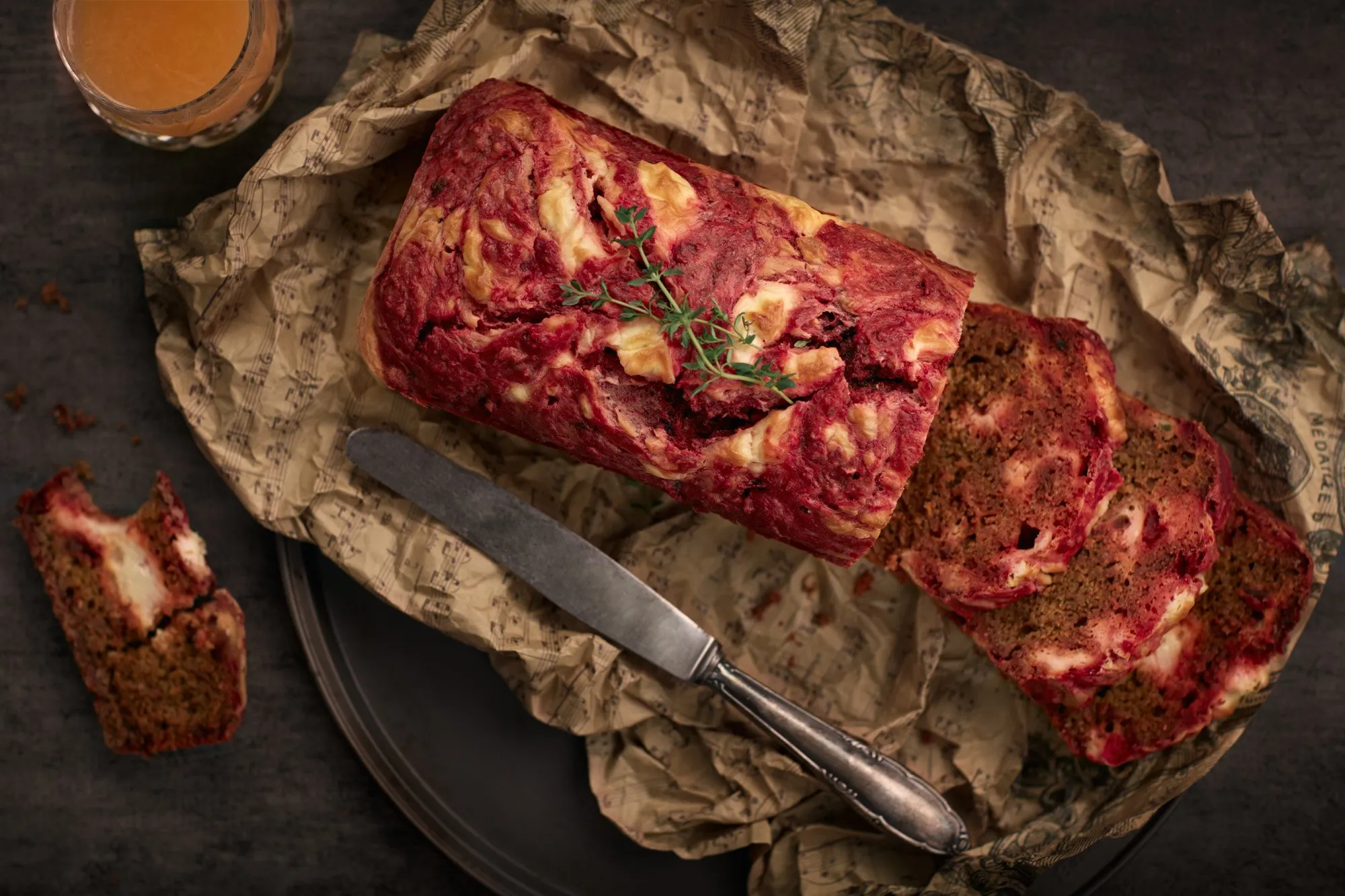 A meatloaf on newspaper, accompanied by a knife, with a few slices and a drink in a glass, on a dark background.