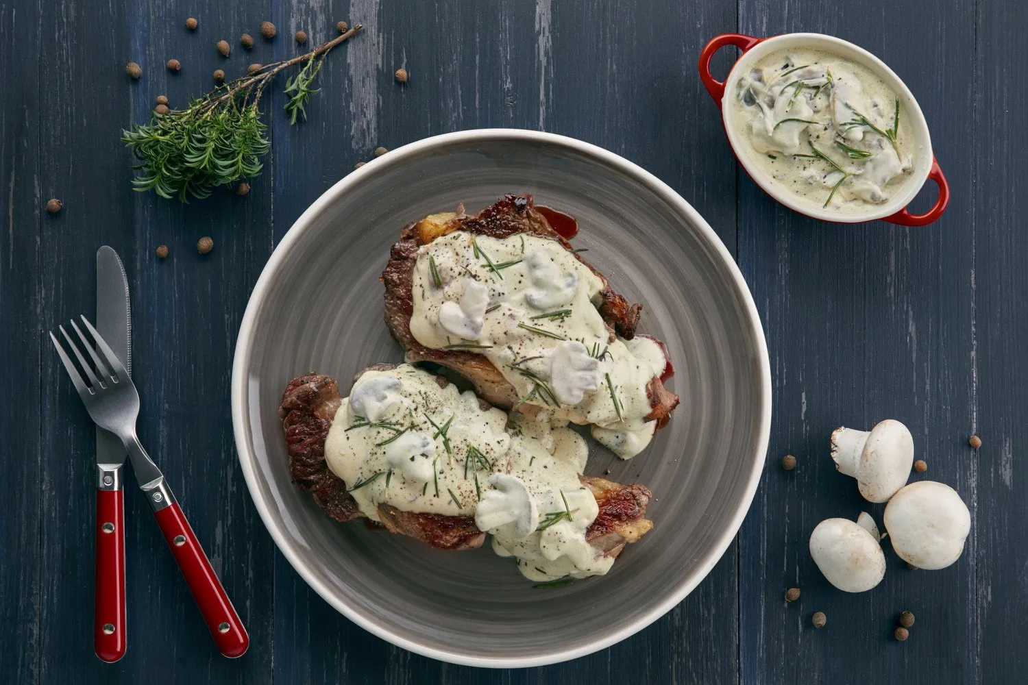 Meat with béchamel sauce and herbs, served with white mushrooms and black peppercorns on a blue wooden table.
