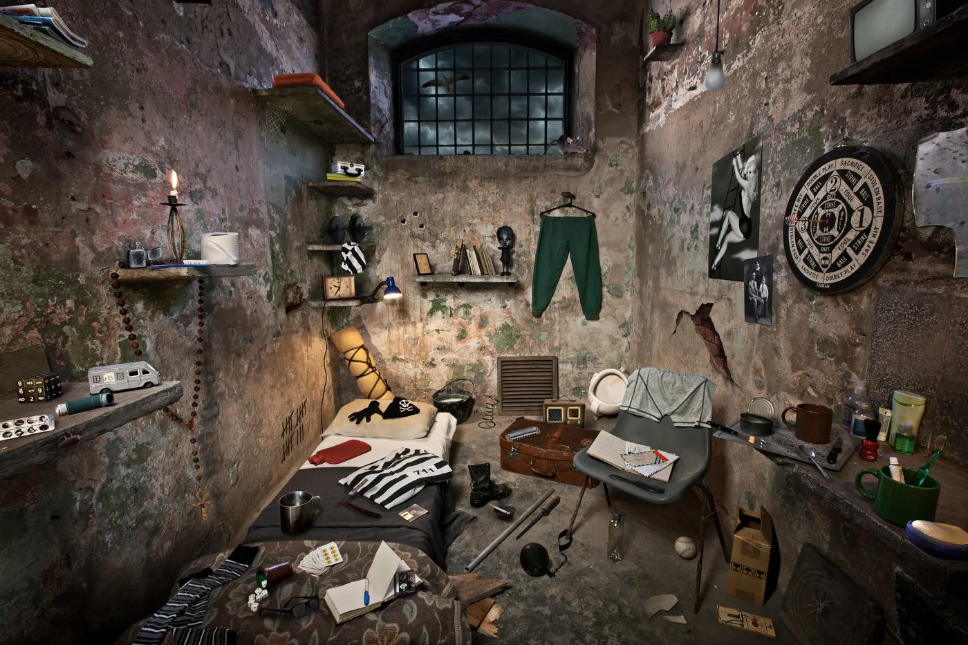 A cluttered room with exposed brick walls and scattered objects, including a bed, a chair, shelves with books and objects, hanging clothes, and various decorations on the walls, in a worn, neglected-looking setting