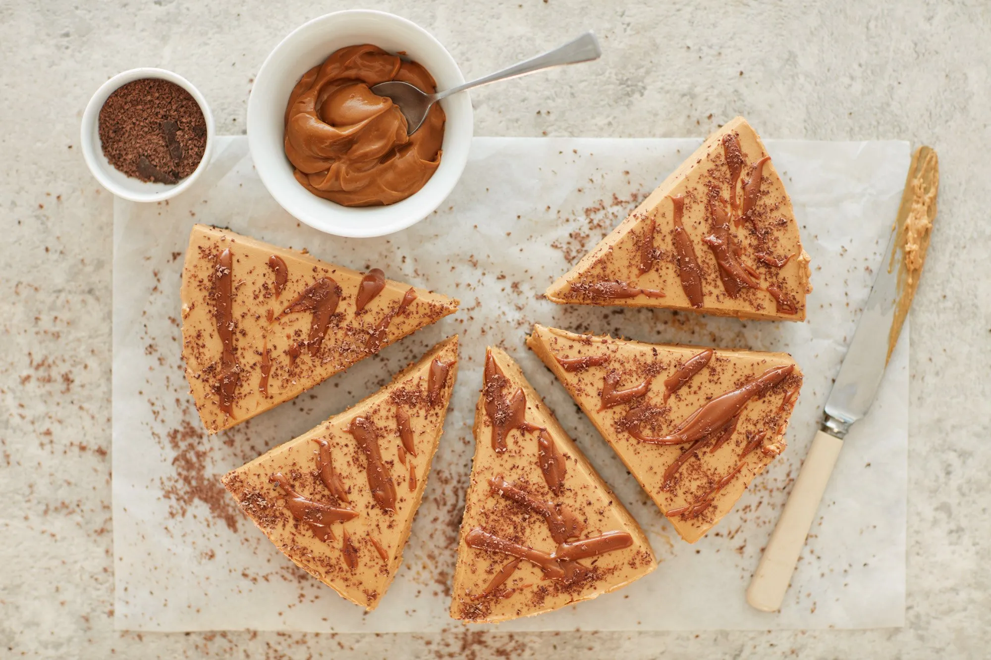 Six slices of caramel cake with chocolate and cocoa powder on wax paper, with chocolate syrup on top, and two containers of dulce de leche cream and cocoa powder alongside.