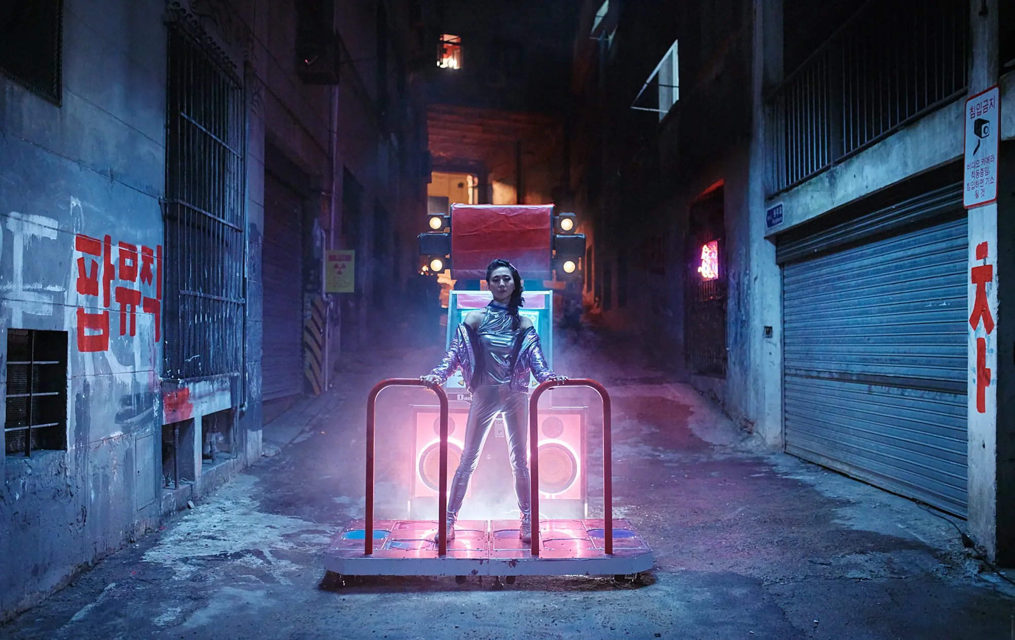 A woman dressed in futuristic clothing standing on a platform in a dark alley with neon and smoke, with speakers and a loading structure in front of her.