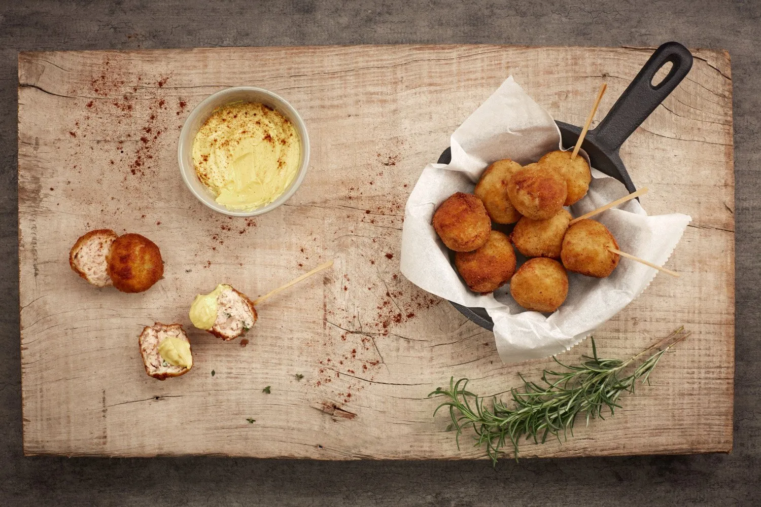 A rustic wooden table with a bowl of yellow sauce, croquettes on skewers, and a sprig of rosemary.