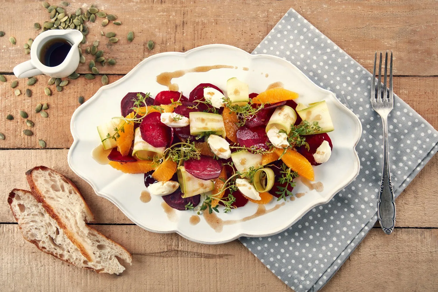 Salad of beet, orange, cucumber, and cheese on a white oval plate, with bread on the side, on a wooden table with a blue linen napkin with white dots and a fork on the right.