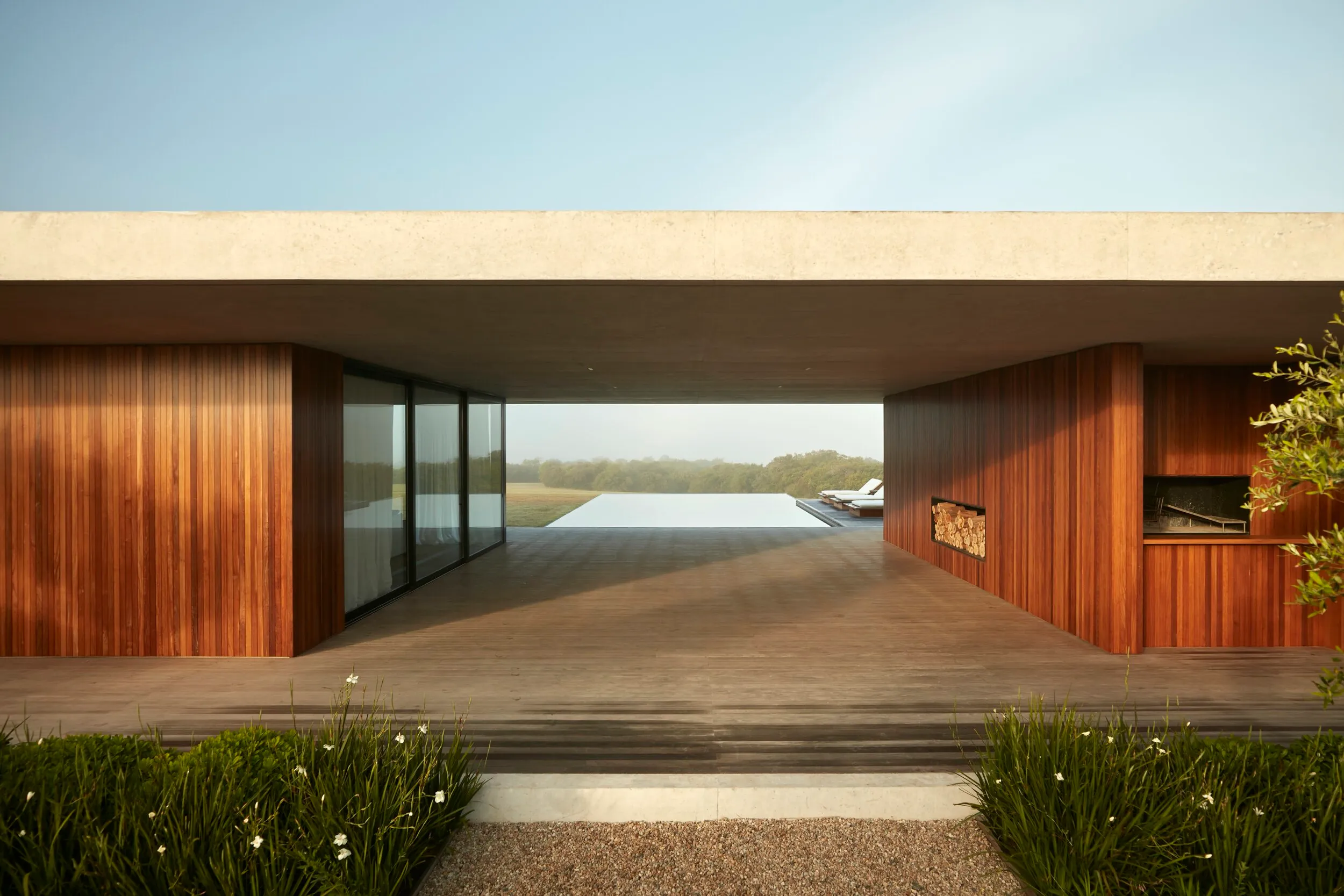 Front view of a modern house with a concrete structure and wooden walls, a pool and chairs in the background, and greenery in the foreground.