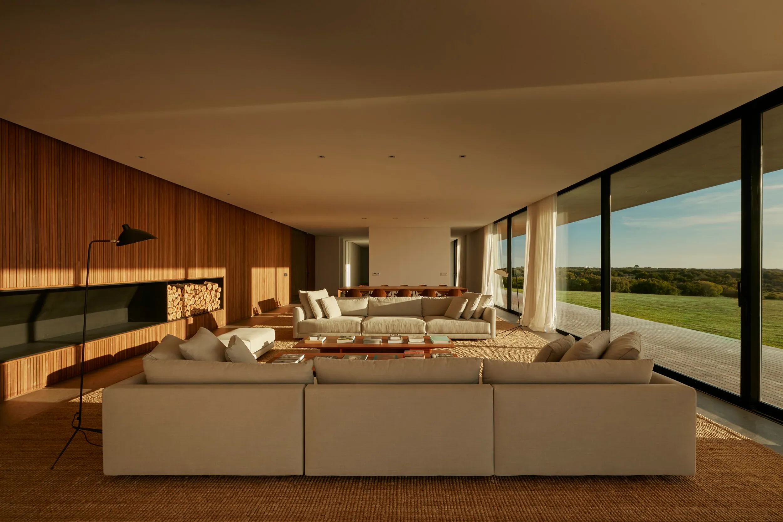 Modern living room with large windows, beige armchairs, a wooden coffee table, a black floor lamp, a wooden wall, and a view of a green field.