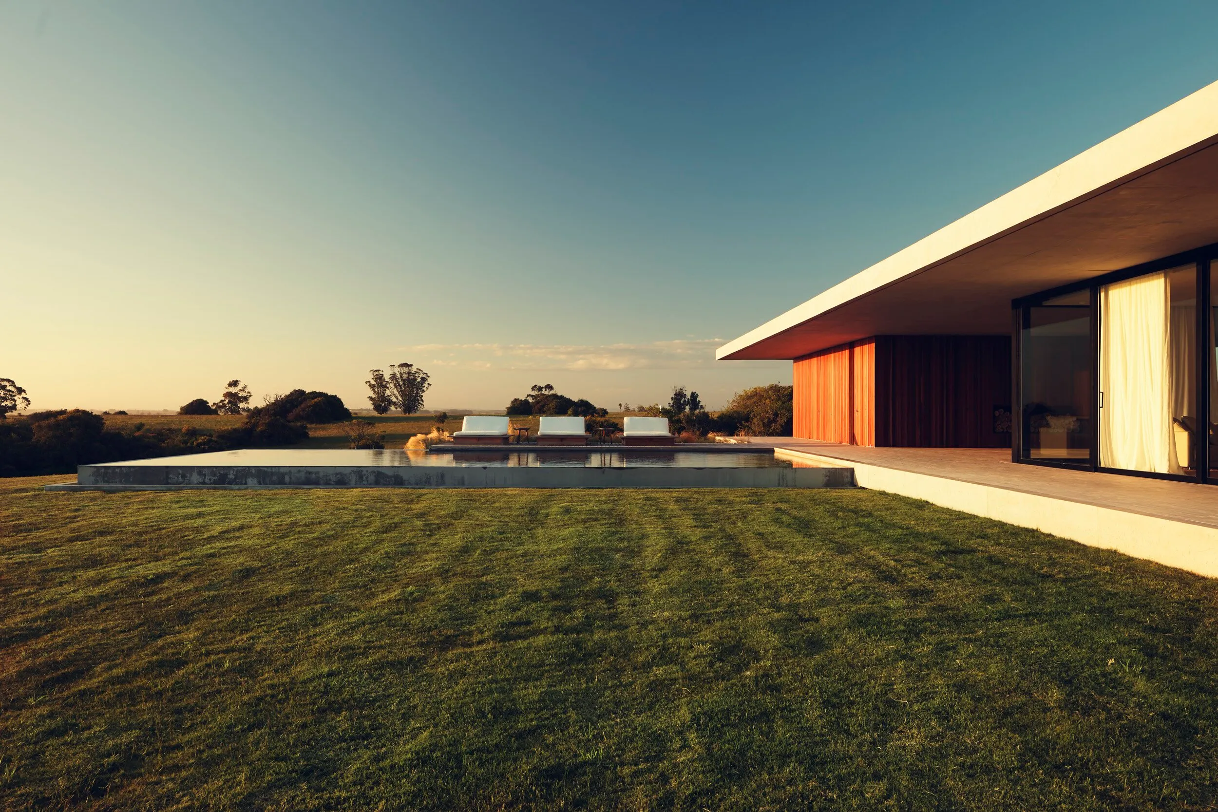 Single-story modern house with a wooden facade and large windows, a pool in the garden and chairs at its edge, in a rural setting with grass and trees