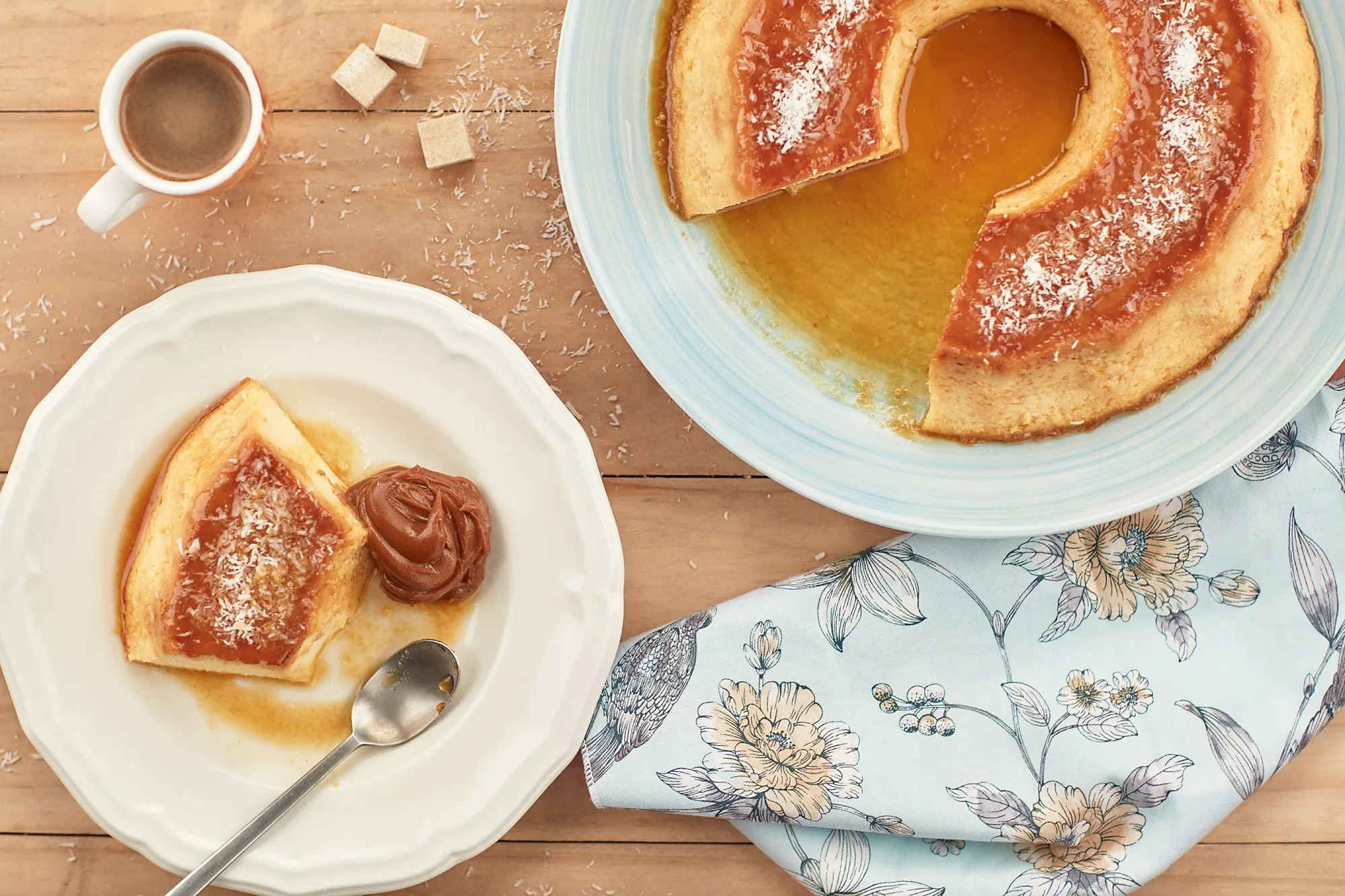A caramel flan on a plate, served with a cut slice and a portion of dulce de leche. There is also a cup of coffee and some sugar cubes on a wooden table set with a floral-patterned tablecloth.