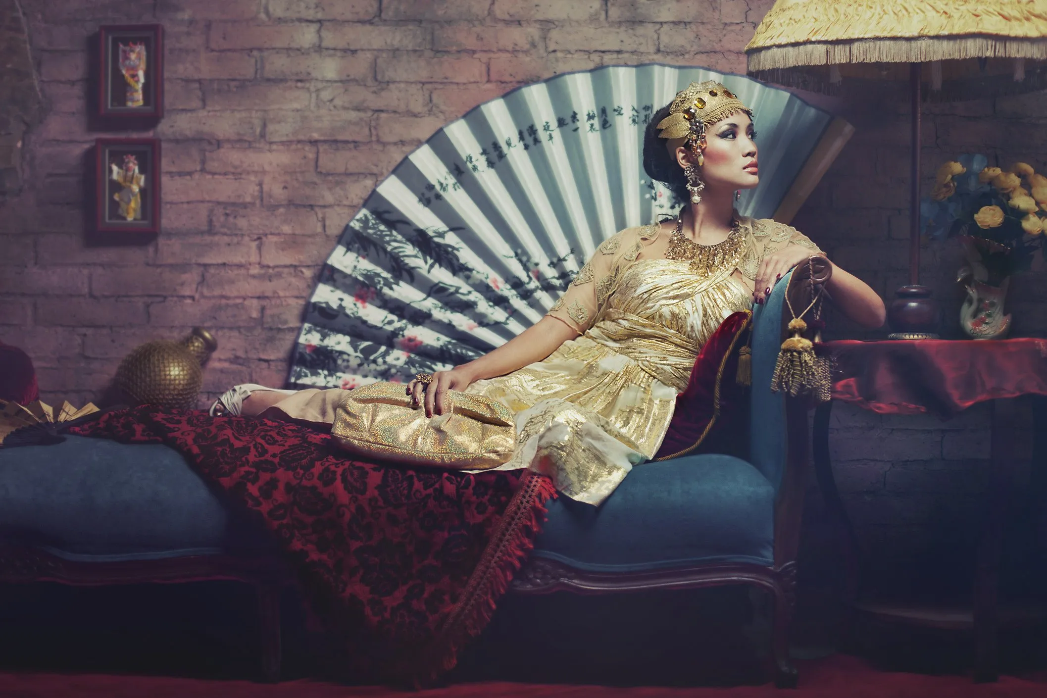 A woman dressed in gold clothing and elaborate accessories, reclining on a blue sofa, with a brick wall and a decorative fan behind her.