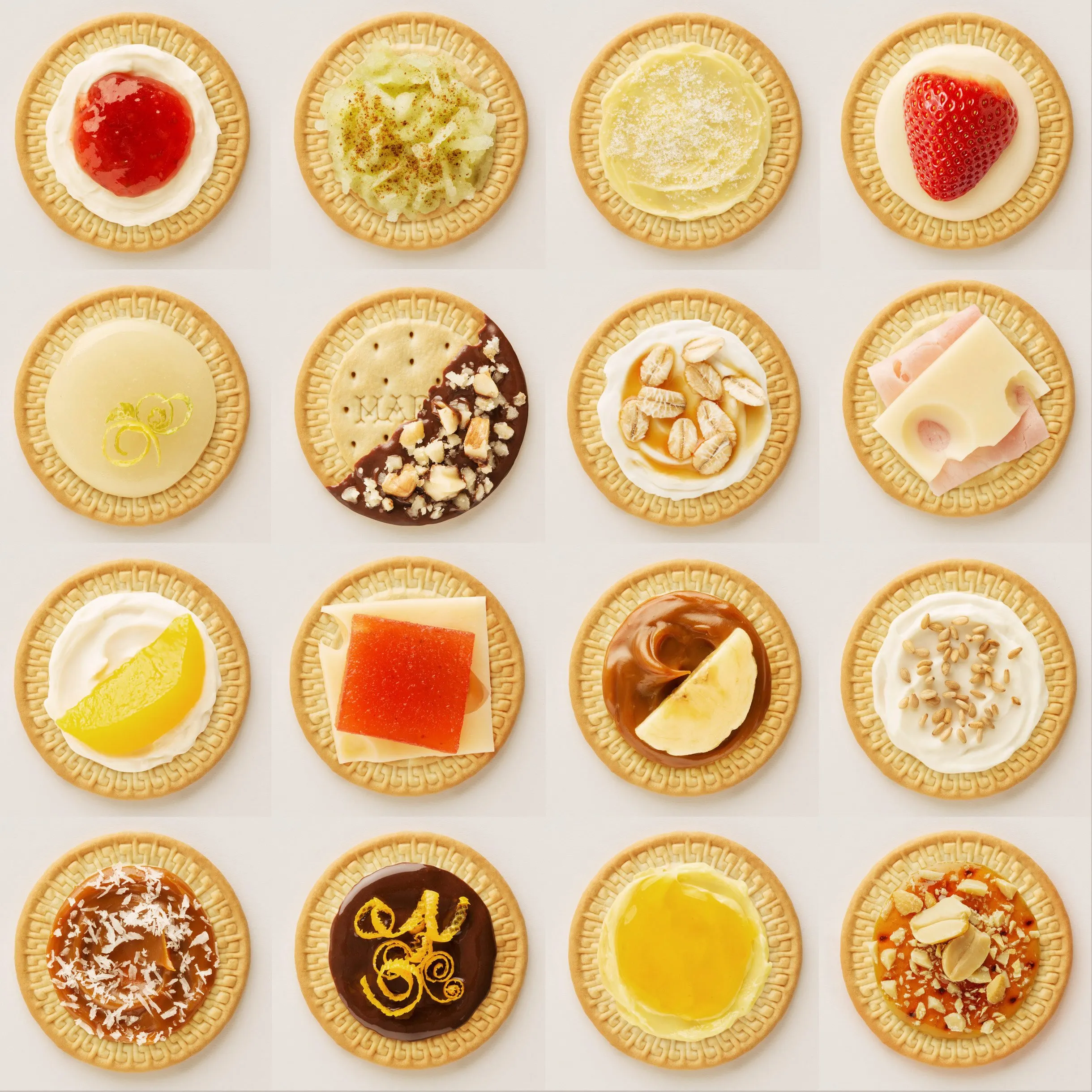 An arrangement of fifteen cookies decorated with different coatings and toppings, including jam, cream, fruit, and chocolate.
