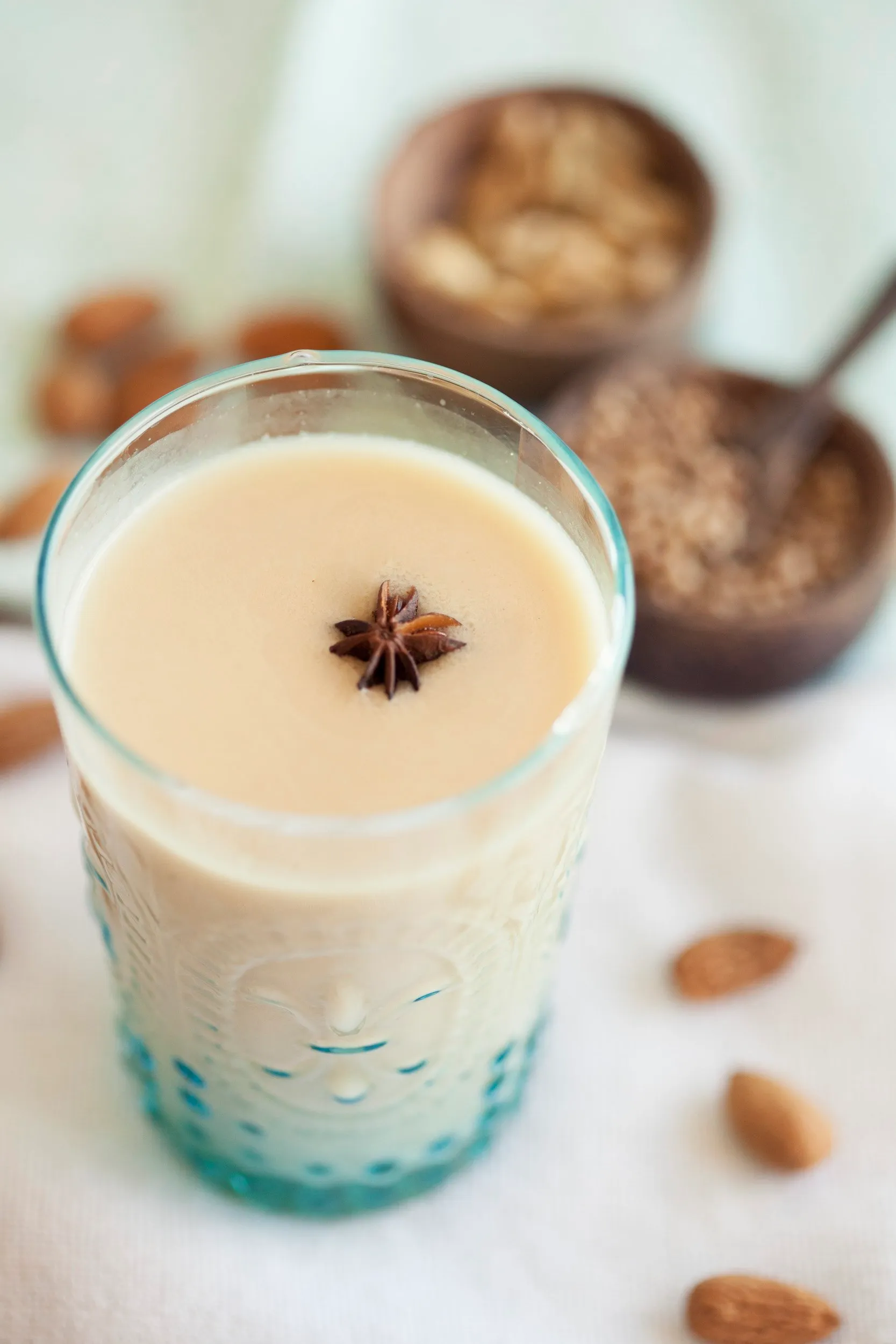 A glass of almond milk with a star anise on the surface, and in the background containers of almonds, cocoa powder, and sugar, on a white table.