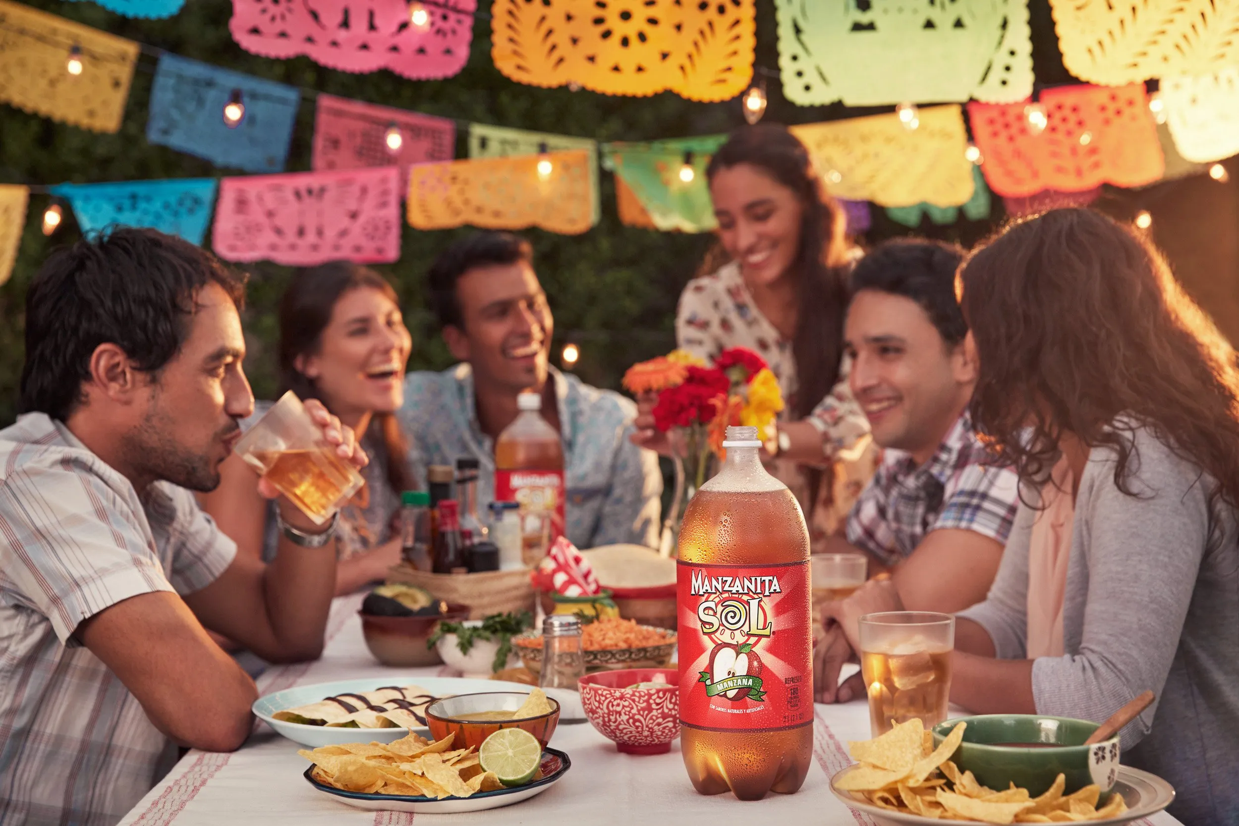 A group of people enjoying an outdoor meal, with colorful decorations and a bottle of Manzanita Sol in the foreground.