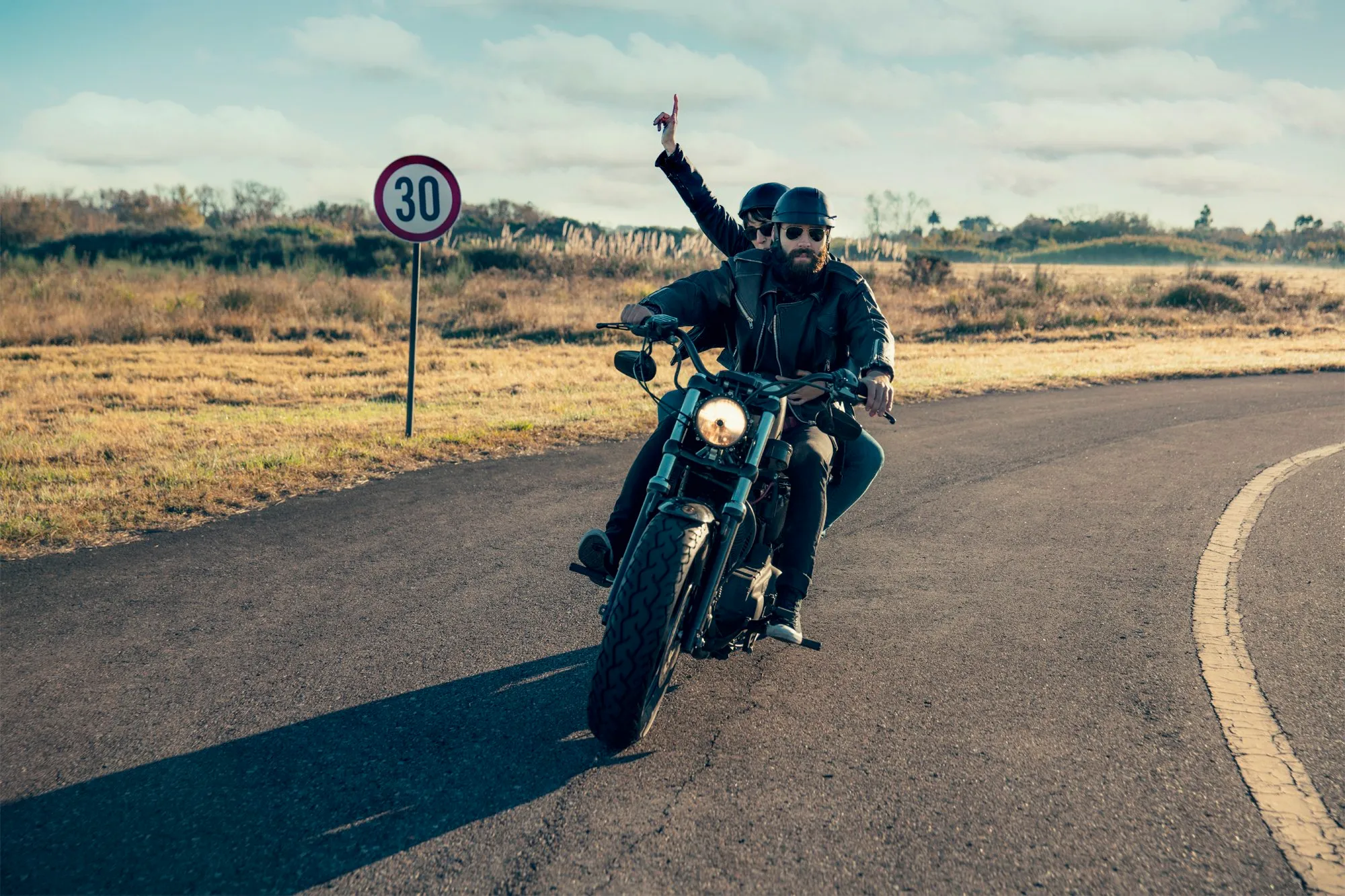 Two motorcyclists wearing helmets, one in front and one behind, riding a motorcycle along a curving road through the countryside, with a 30 km/h speed limit sign.