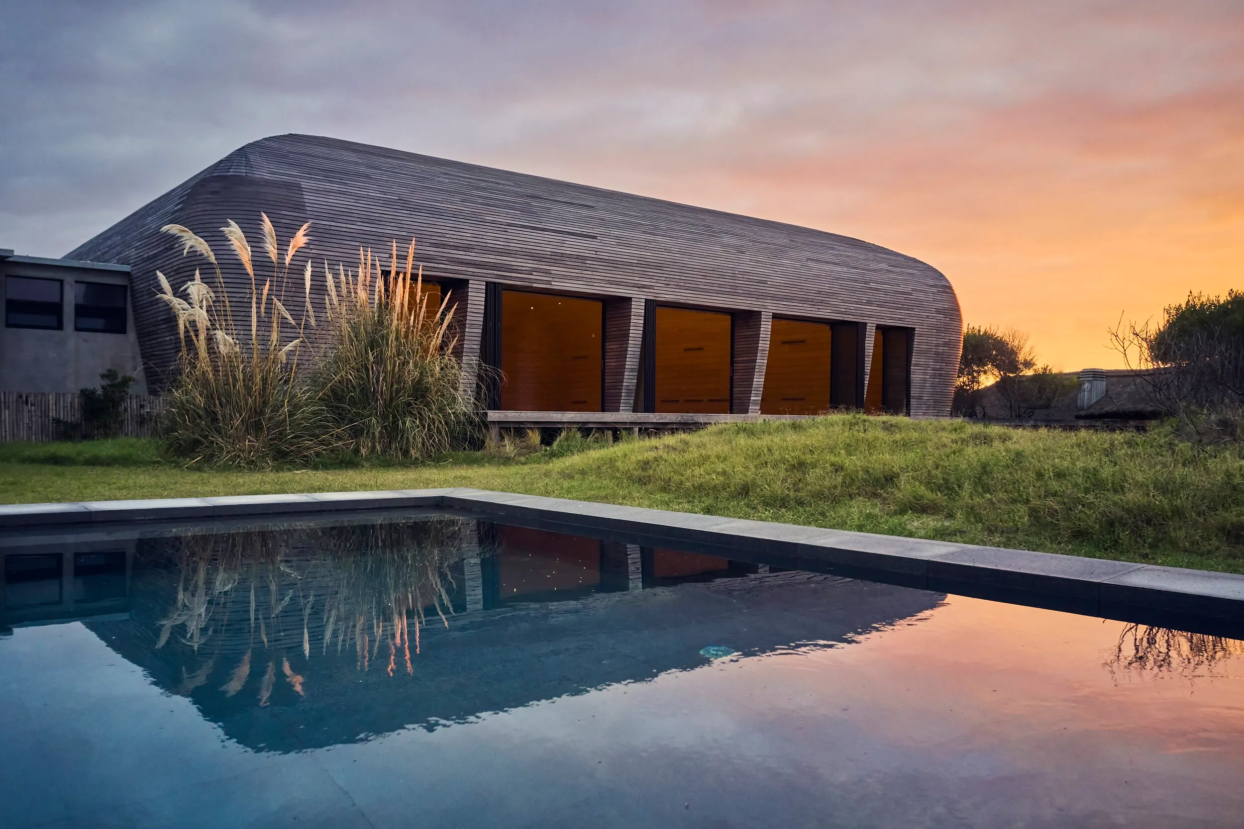 Modern house with a curved design and large windows, next to a pool and surrounded by grass, at sunset under a cloudy sky with warm tones.