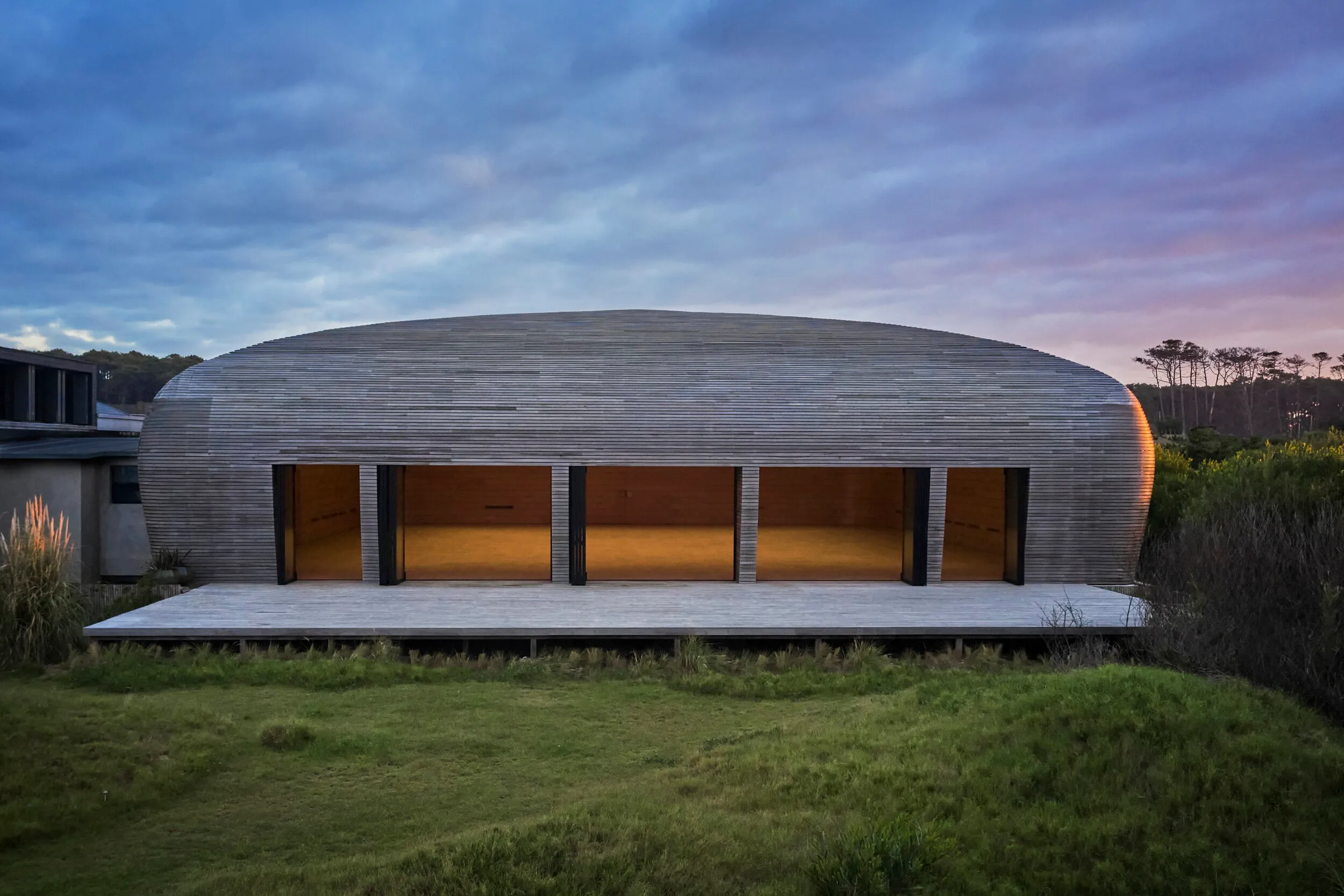 Modern building with a wooden dome, large windows, and a terrace in a natural setting at sunset.