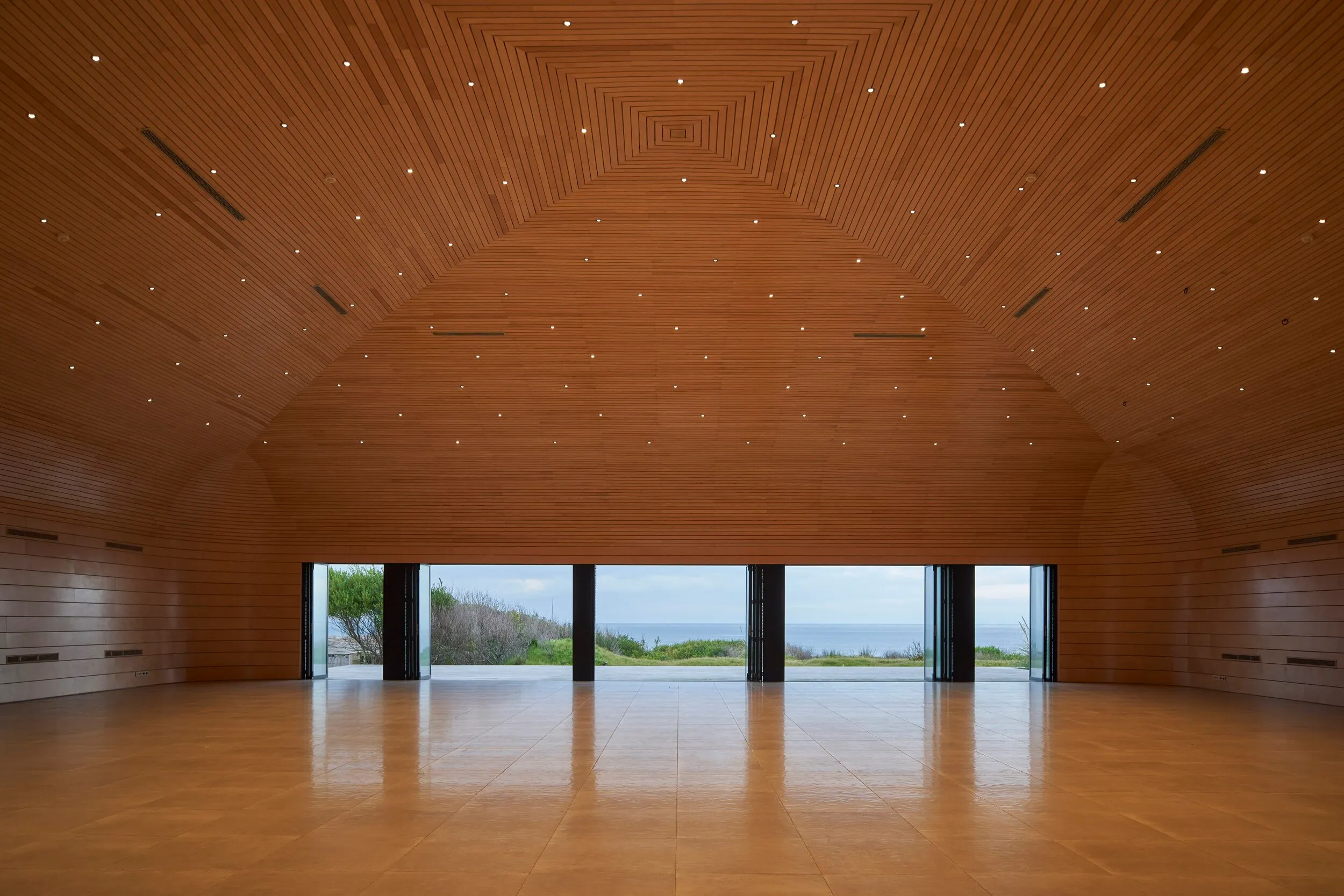 Large room with wooden walls and ceiling, open windows revealing a sea view and a landscape with trees.
