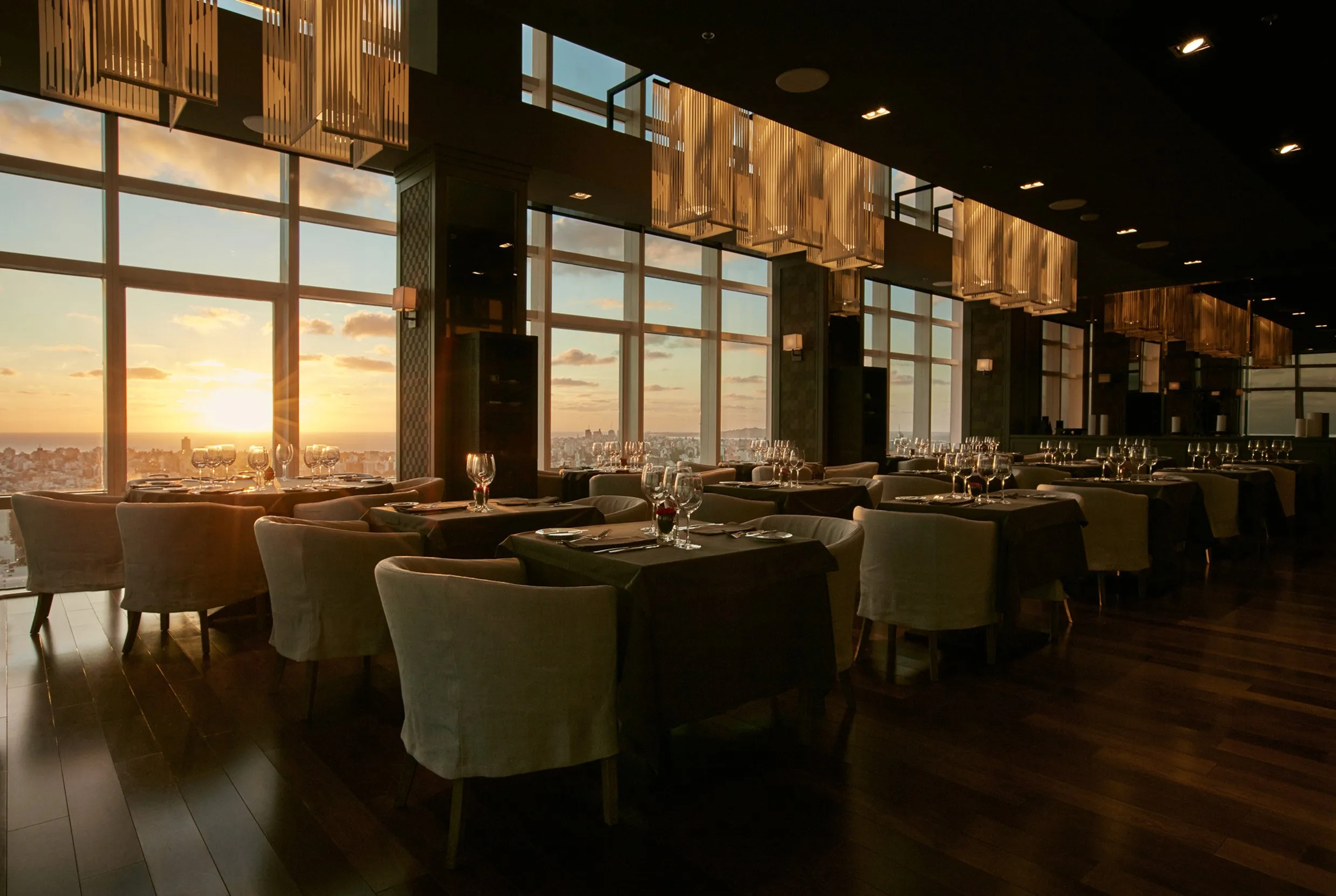 Elegant restaurant with a sunset view, tables set with glassware and cutlery, in a dark setting with large windows and modern decor.