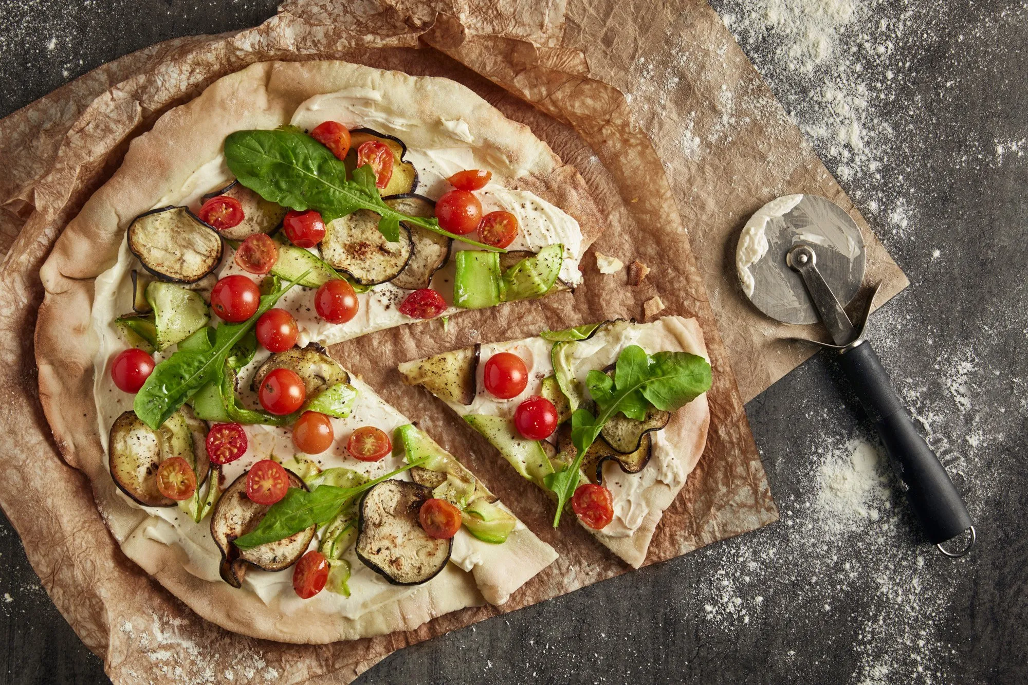 An unbaked pizza with toppings like cherry tomato, eggplant, zucchini, and basil leaves on baking paper.