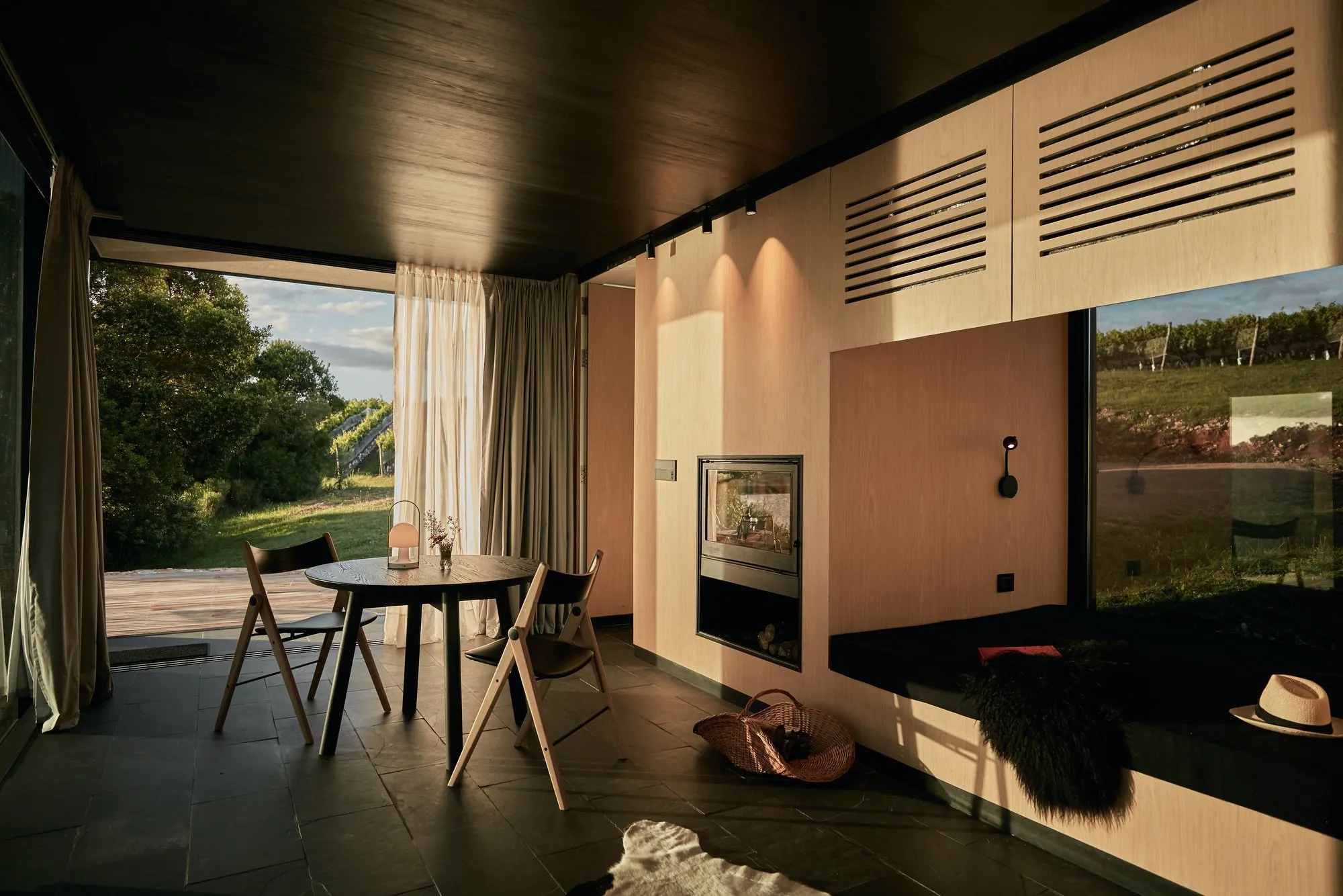 Interior of a modern room with a view of a natural landscape, a round table with chairs, wooden walls, a fireplace, white curtains, and decorative accents