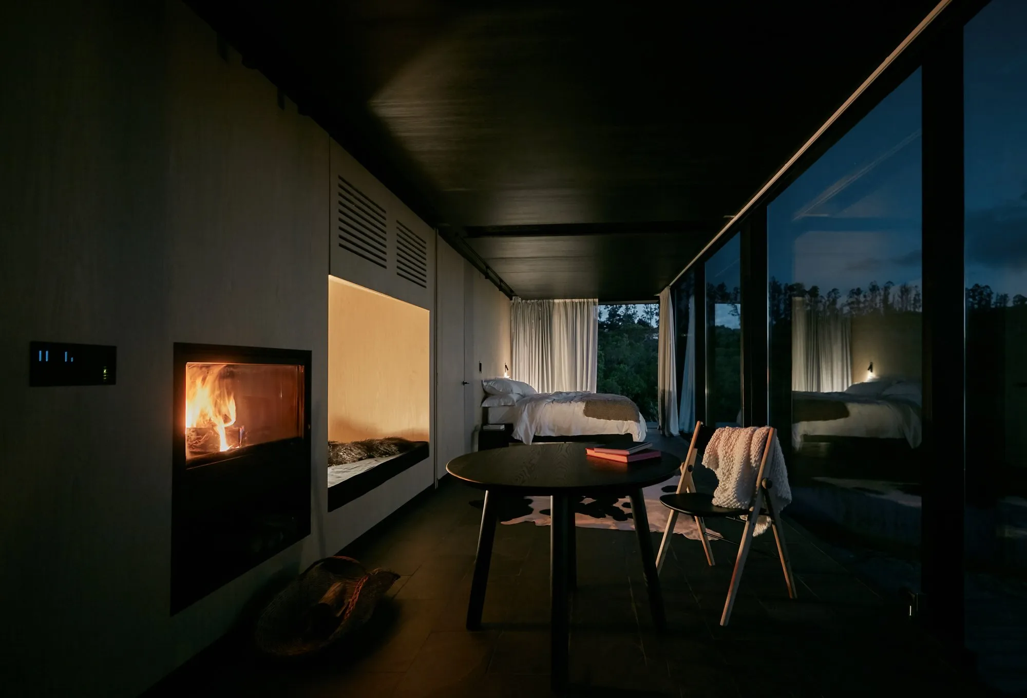 Modern bedroom with a lit fireplace, a large bed, a view of nature through large windows and white curtains, and minimalist decor.