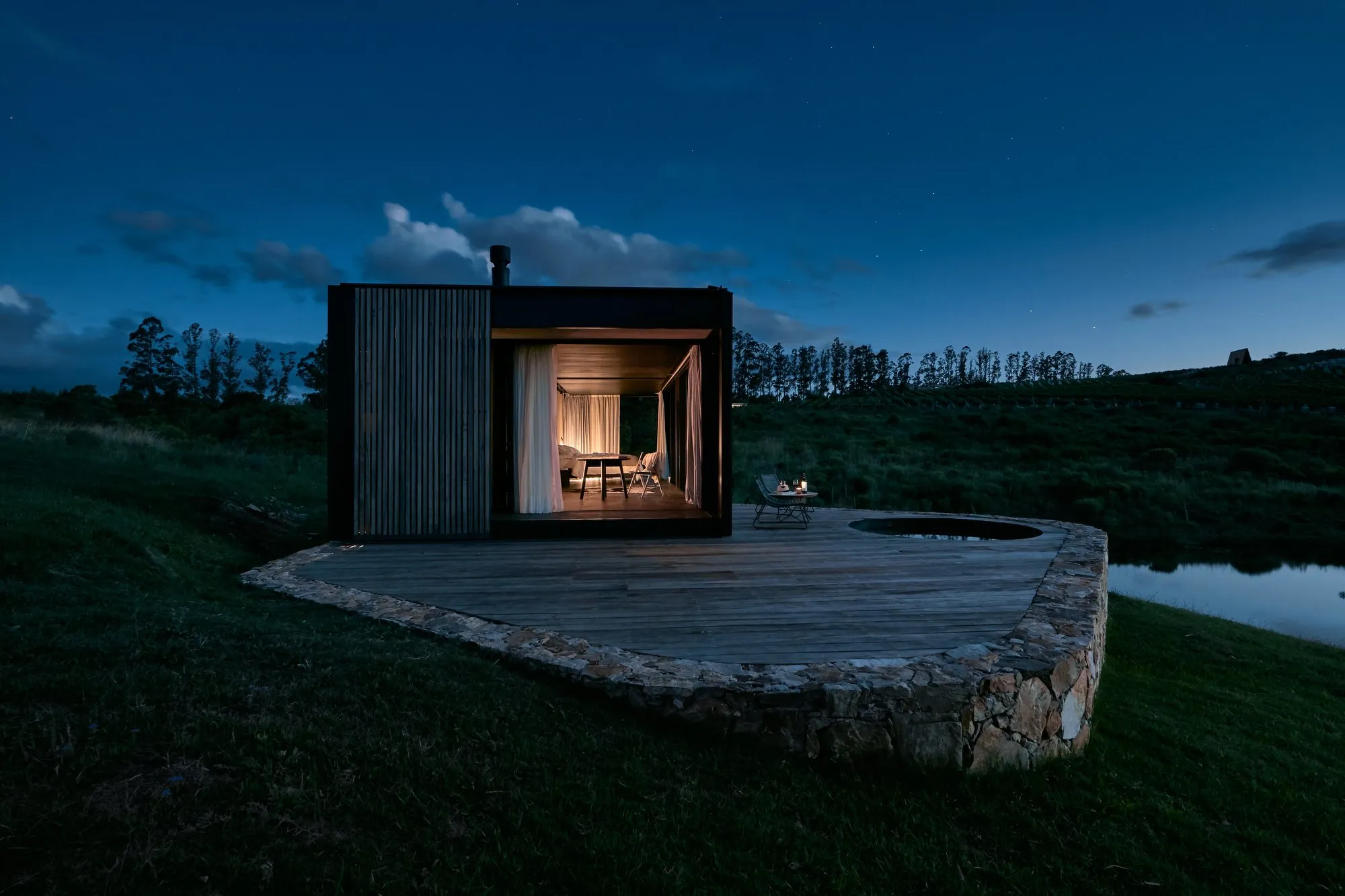 Modern rectangular house with wooden walls and windows with white curtains, in a natural setting with grass and a small body of water beside it, at night under a starry sky.