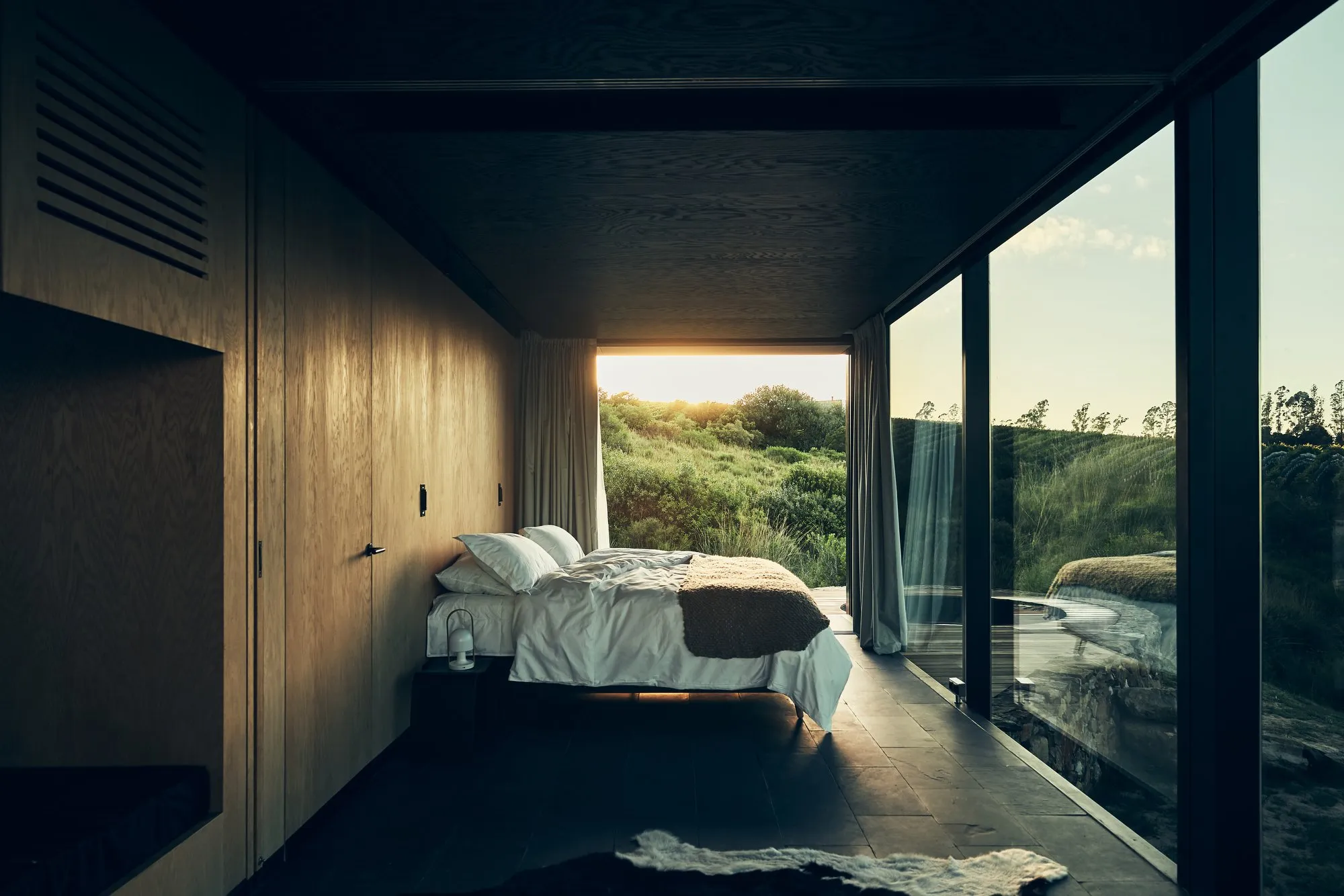 Modern room with a view of nature, a bed, wooden walls, and large windows with white curtains opening onto a green, sunny landscape.