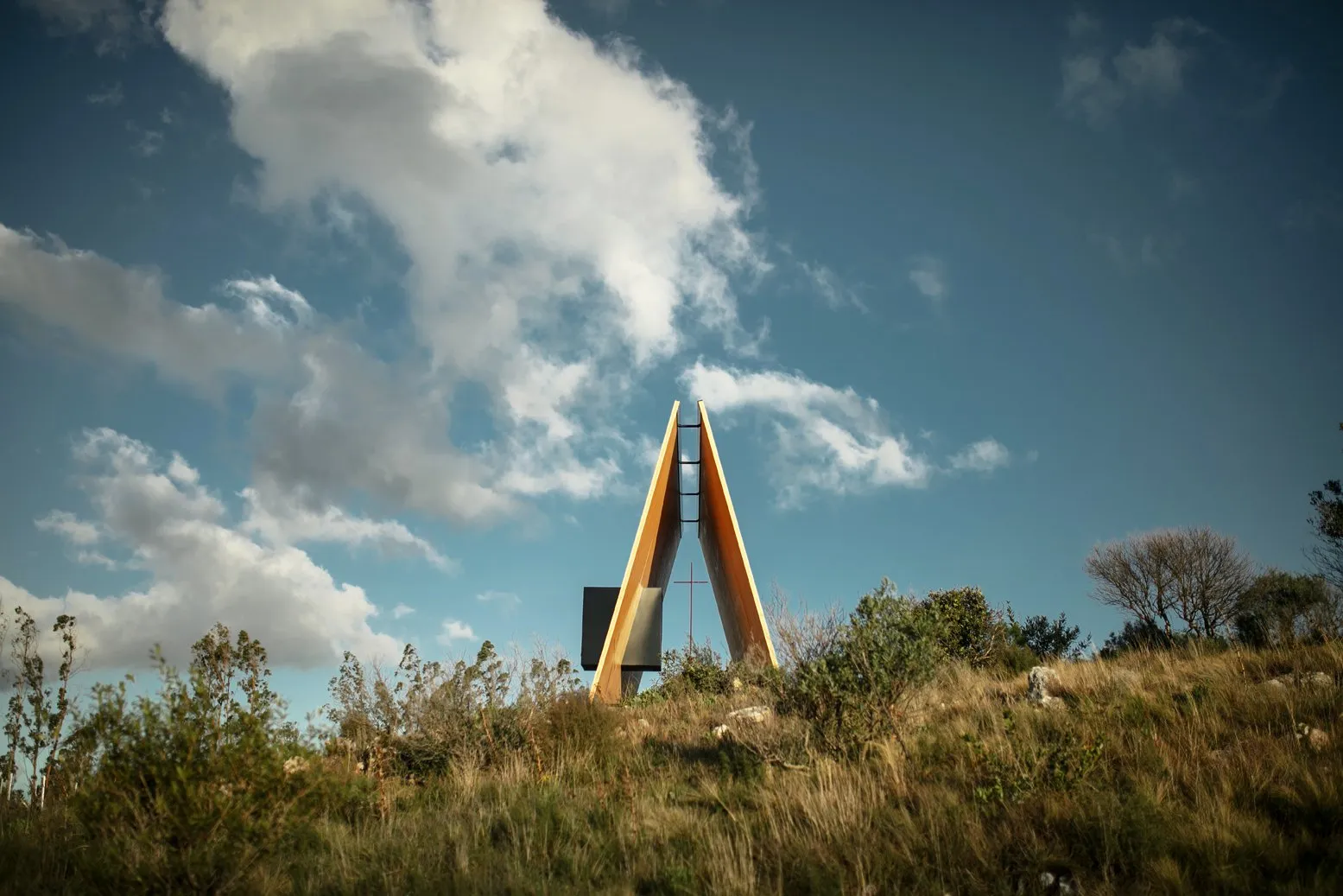 Modern structure on a hill with trees and a cloudy sky behind.