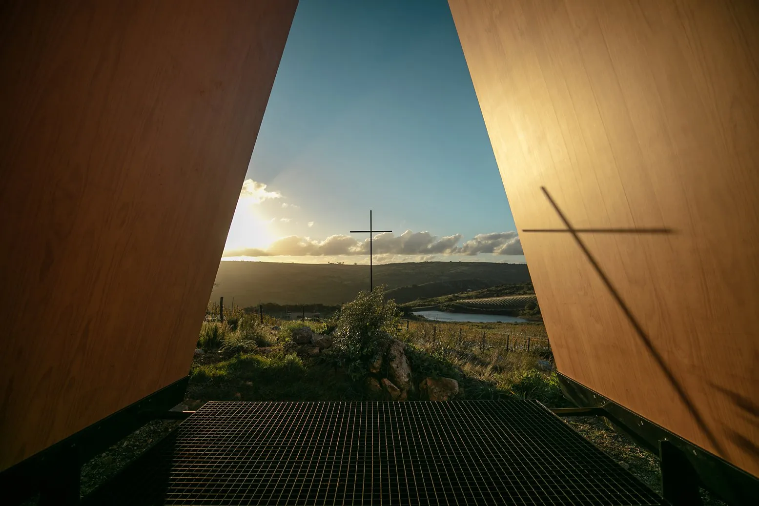 View of a countryside landscape with hills, a river, and a crucifix in the background, seen from a wooden structure with a metal frame in the foreground, at sunset.