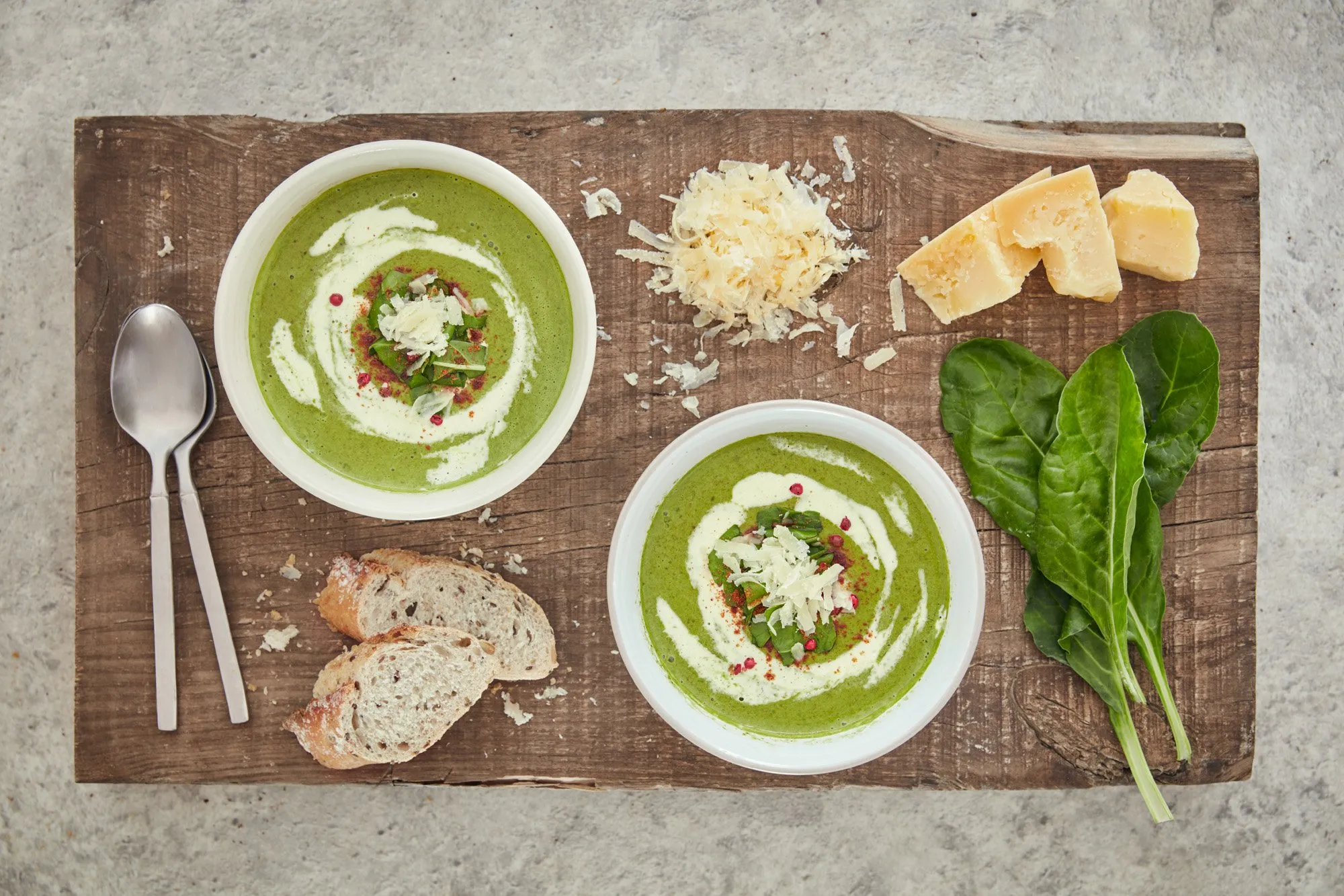 Spinach soup with cream and grated cheese in white bowls, homemade bread, cheese, spinach, and herbs on a wooden board.