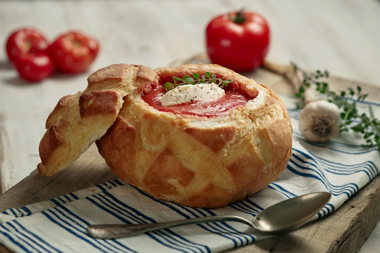 A soup served in a baked bread bowl, with cheese, tomato, and herbs, on a wooden board with a spoon and tomatoes in the background.