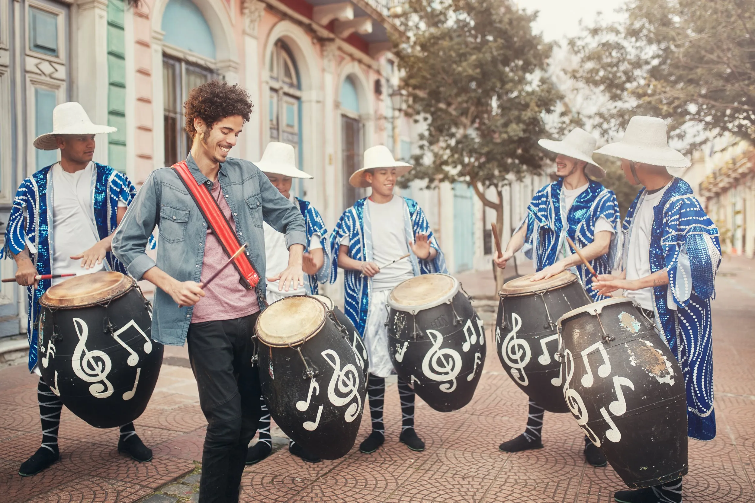 A group of musicians playing drums decorated with musical notes on a street with trees and colorful buildings.
