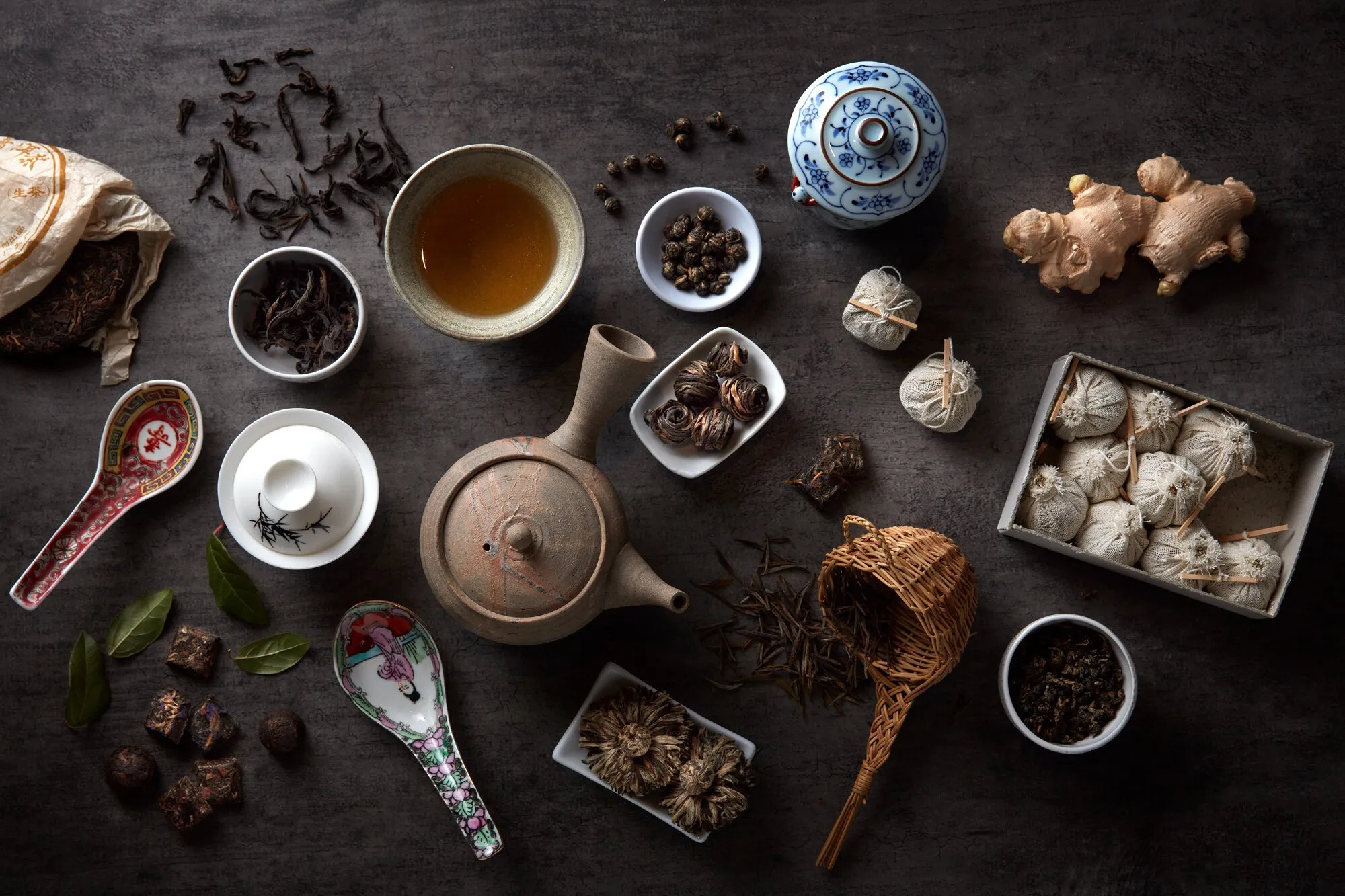 Various ingredients and utensils for preparing Chinese tea, including loose teas, ginger, and traditional accessories on a dark wooden surface.