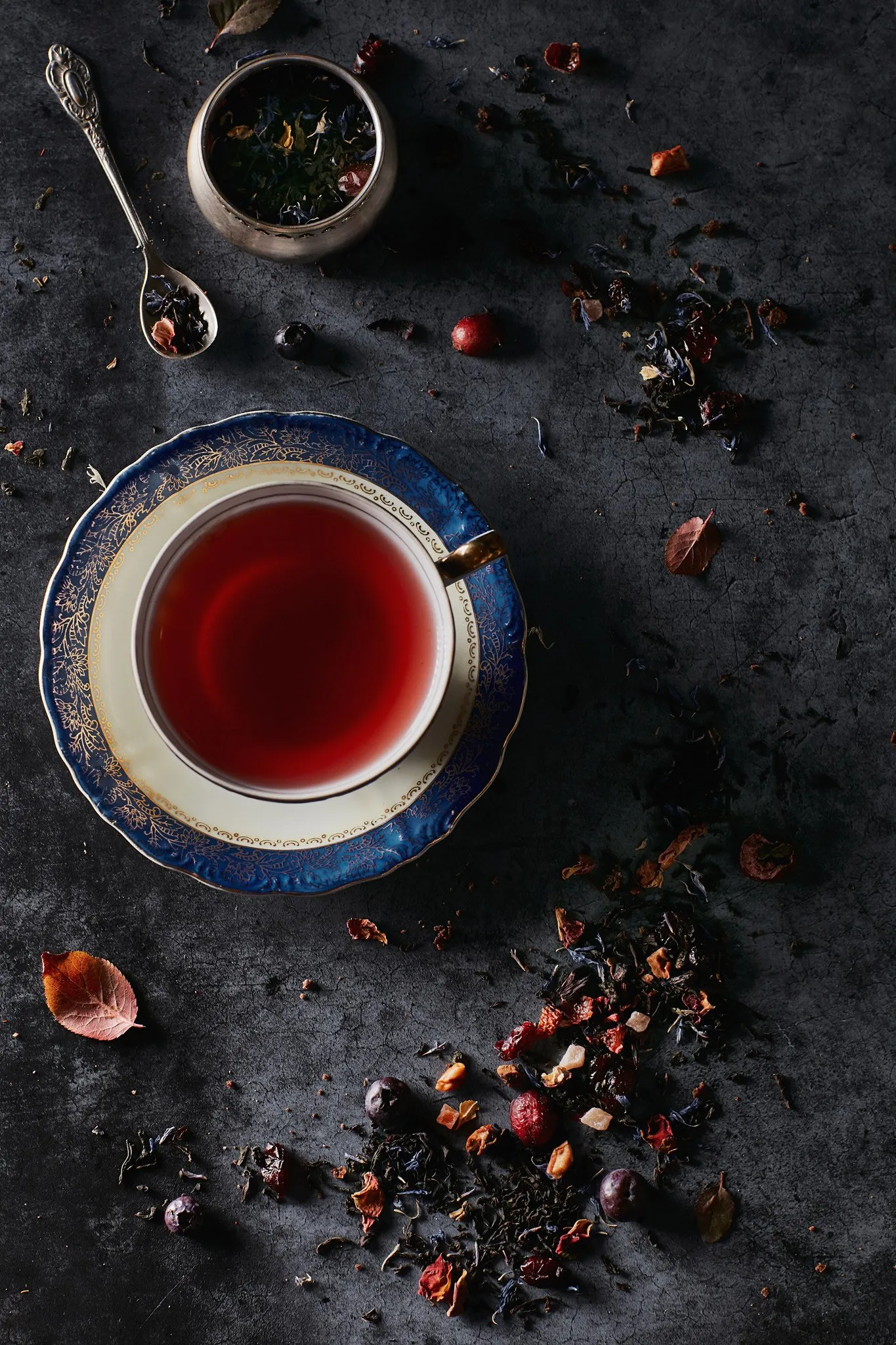 A cup of red tea on a decorative blue and gold saucer, surrounded by dried flowers, leaves, and pieces of fruit that look like blueberries and rose petals, on a dark surface.