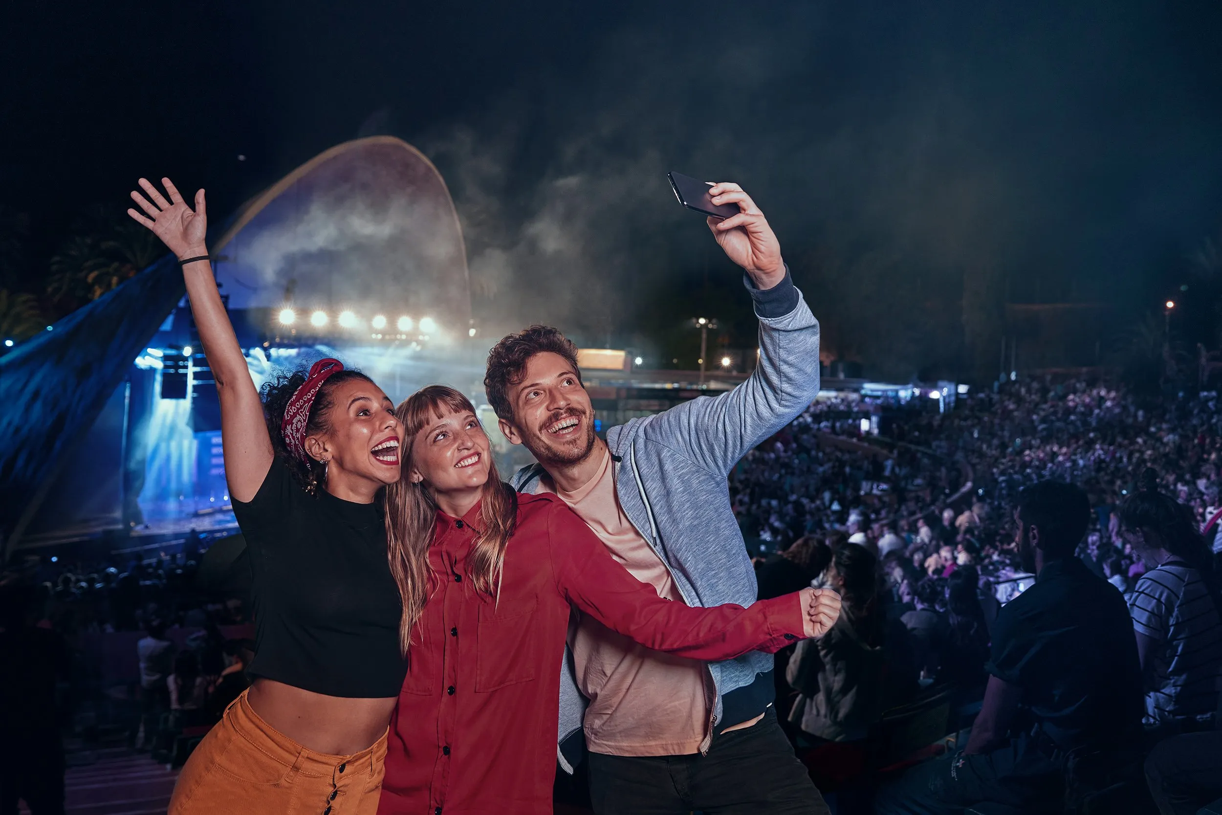 Three happy people taking a selfie at an outdoor concert at night, with an audience and a lit stage in the background.