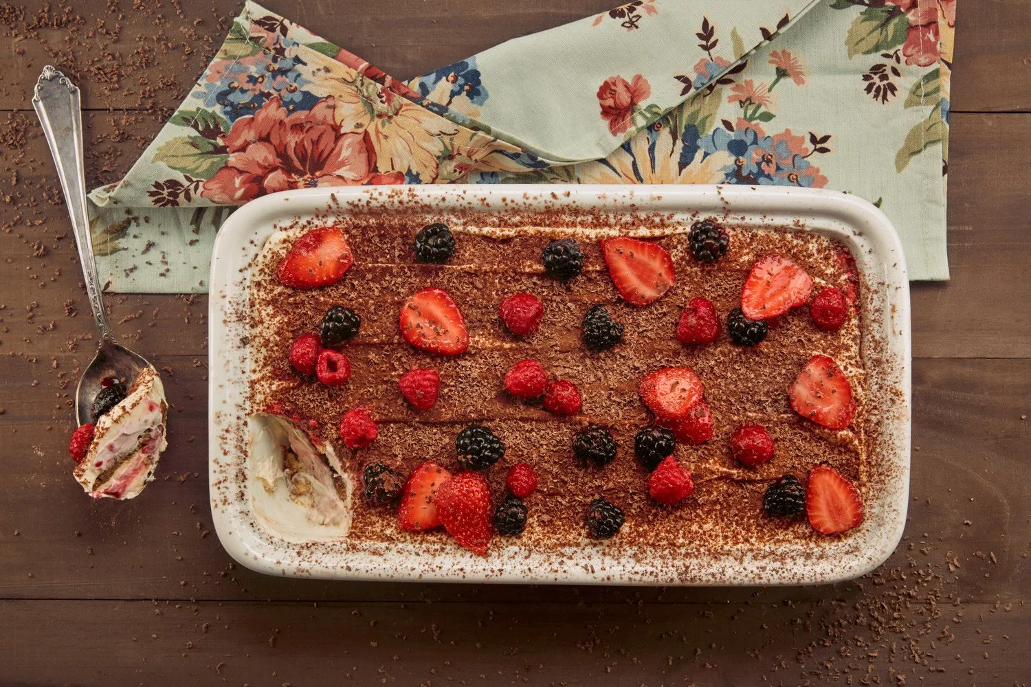 A tiramisu dessert topped with raspberries, strawberries, and blackberries, on a white rectangular plate on a wooden table. A fork with a piece of the dessert and a napkin with a floral design are on the left.