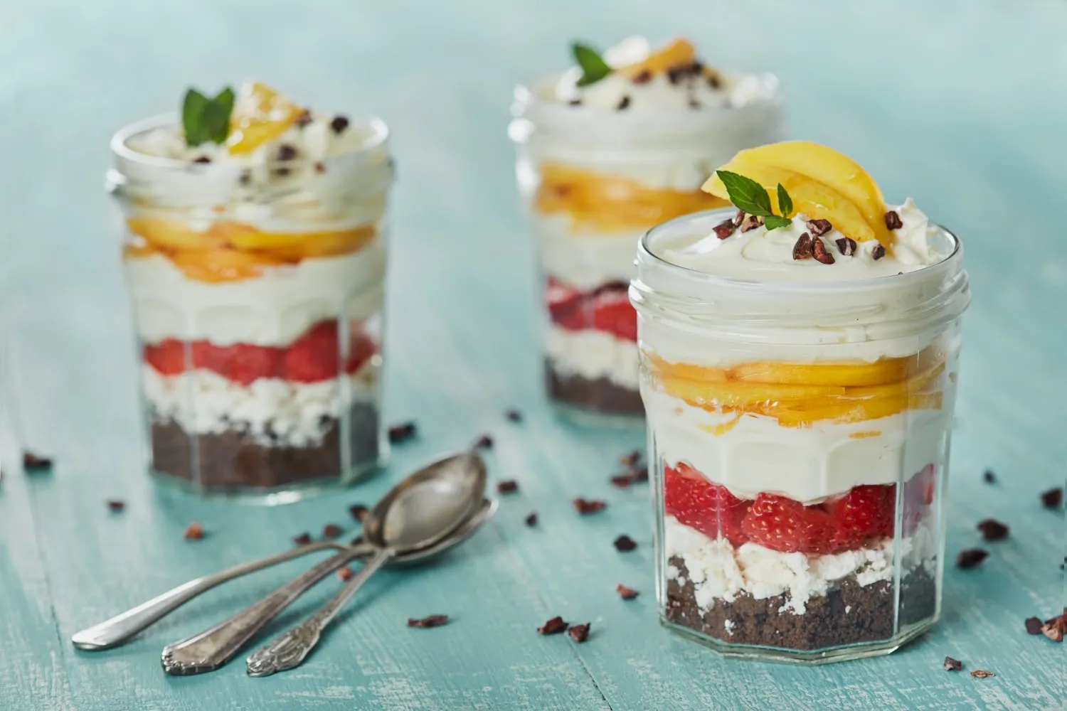 Glasses layered with chocolate cake, cream, strawberries, and mango, decorated with mint and cocoa shavings, on a blue wooden table.