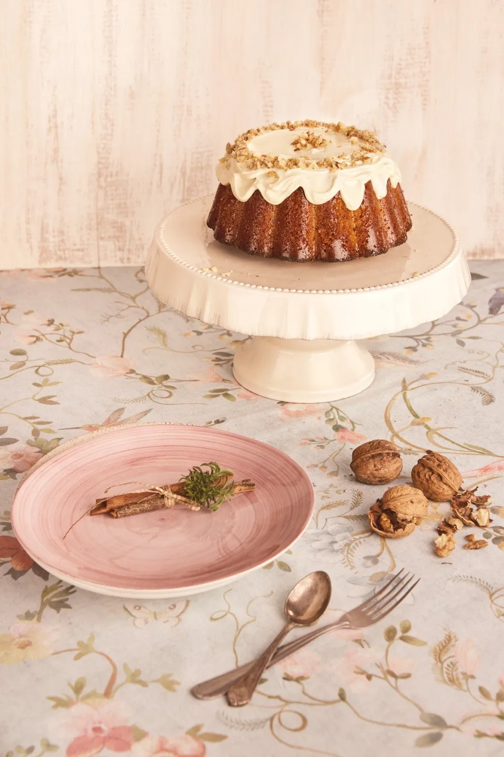 A cake on a stand decorated with walnuts and cream, a pink plate with a sprig of greenery and walnuts on the table, and silver cutlery beside a backdrop of flowers on the table