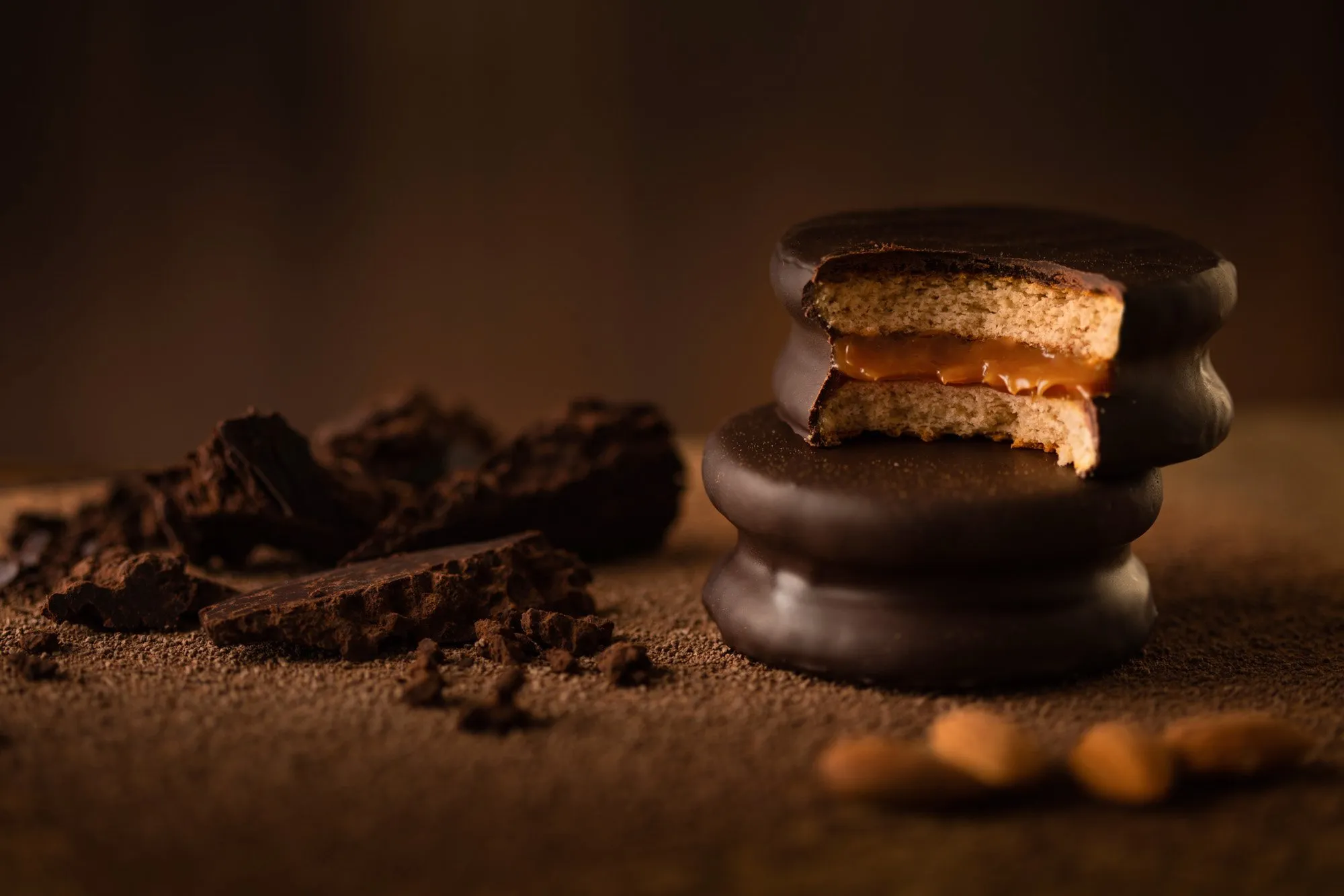 A cookie with a caramel filling, coated in chocolate, on a surface of cocoa powder next to pieces of chocolate