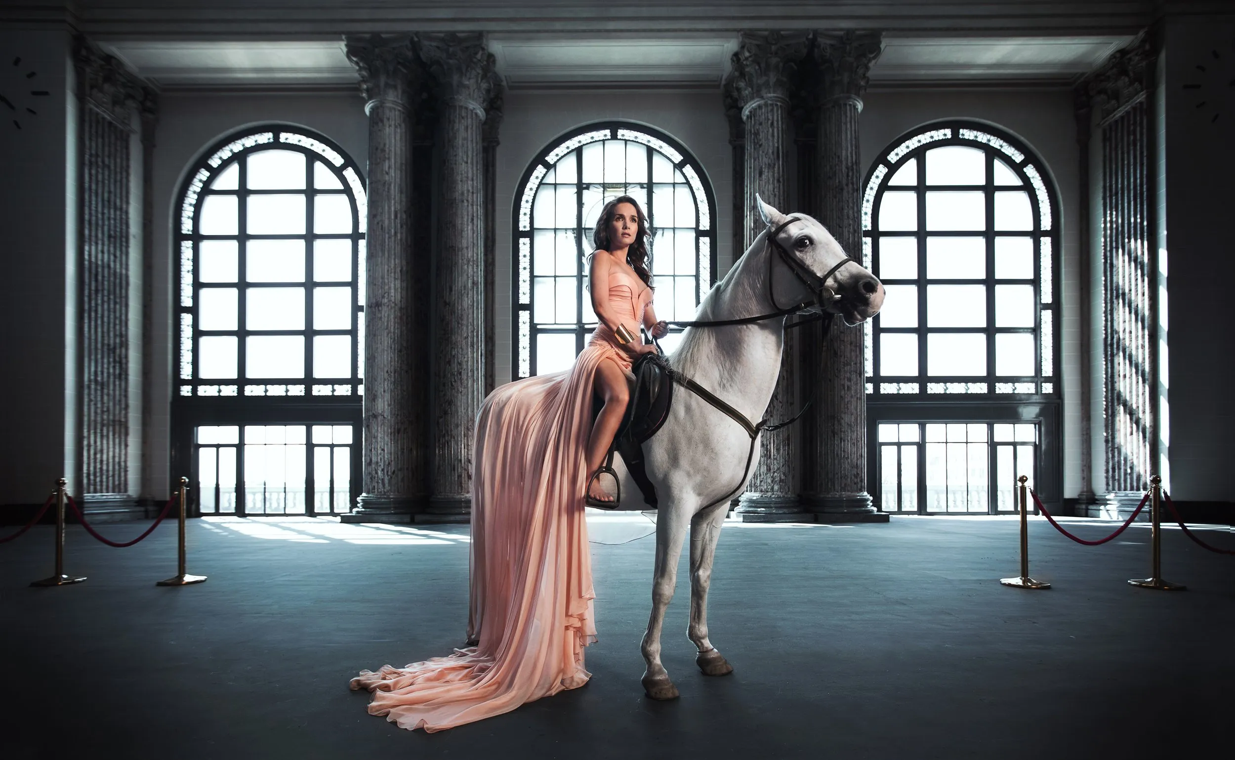 A woman in a long, light pink dress riding a white horse indoors, with stone columns and tall windows