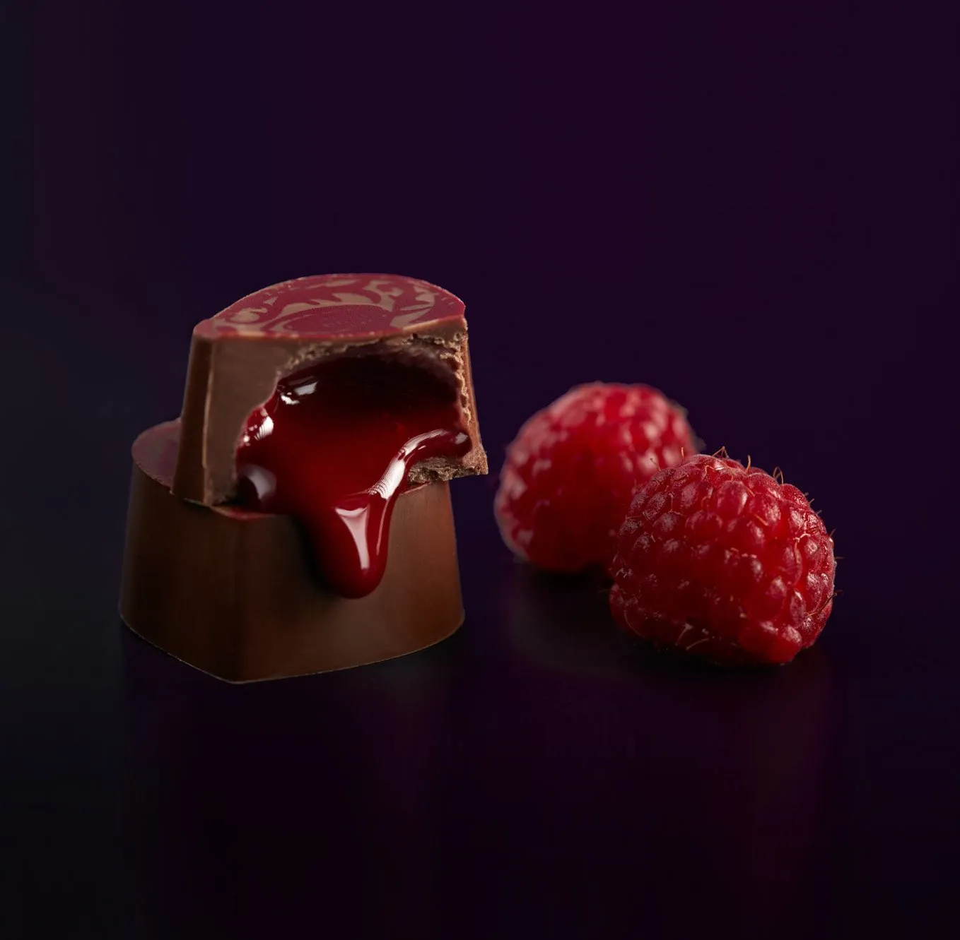 A chocolate filled with raspberry sauce next to two fresh raspberries.