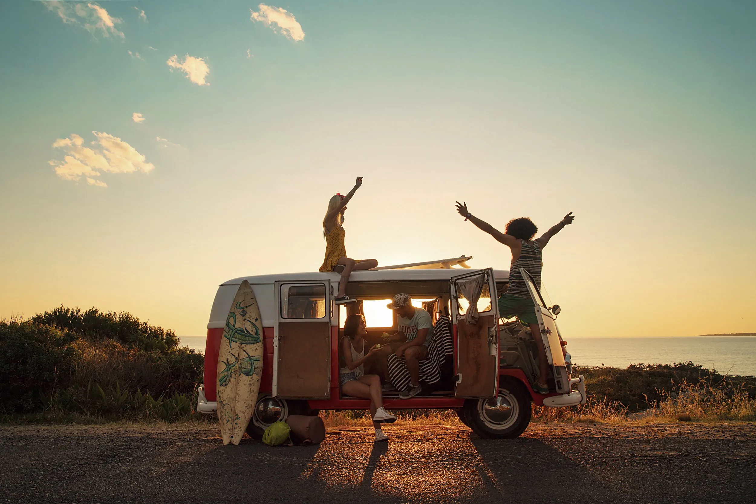 A group of friends having fun in a van with a sea view at sunset, with a girl on the roof and others inside, in a relaxed, cheerful mood.