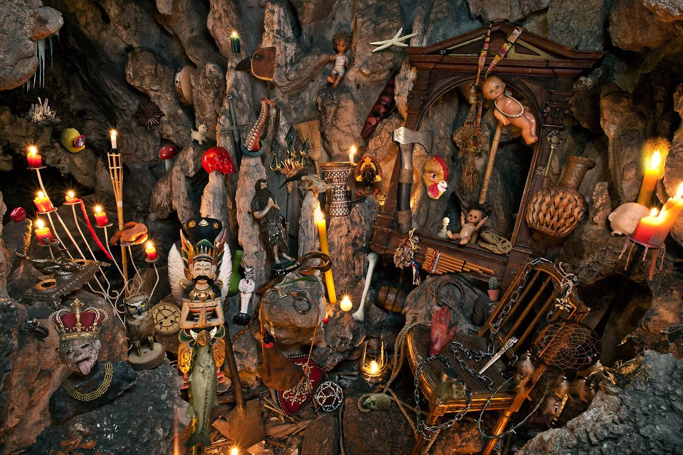 An altar with ritual and decorative objects, including lit candles, masks, figures, weapons, chains, a hanging fish, and various other elements in a stone setting.