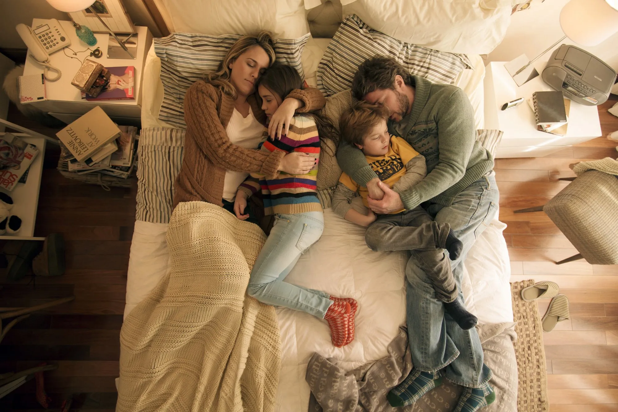 A family of five asleep in a bed, with a small child hugging their mother and another small child hugging their father.