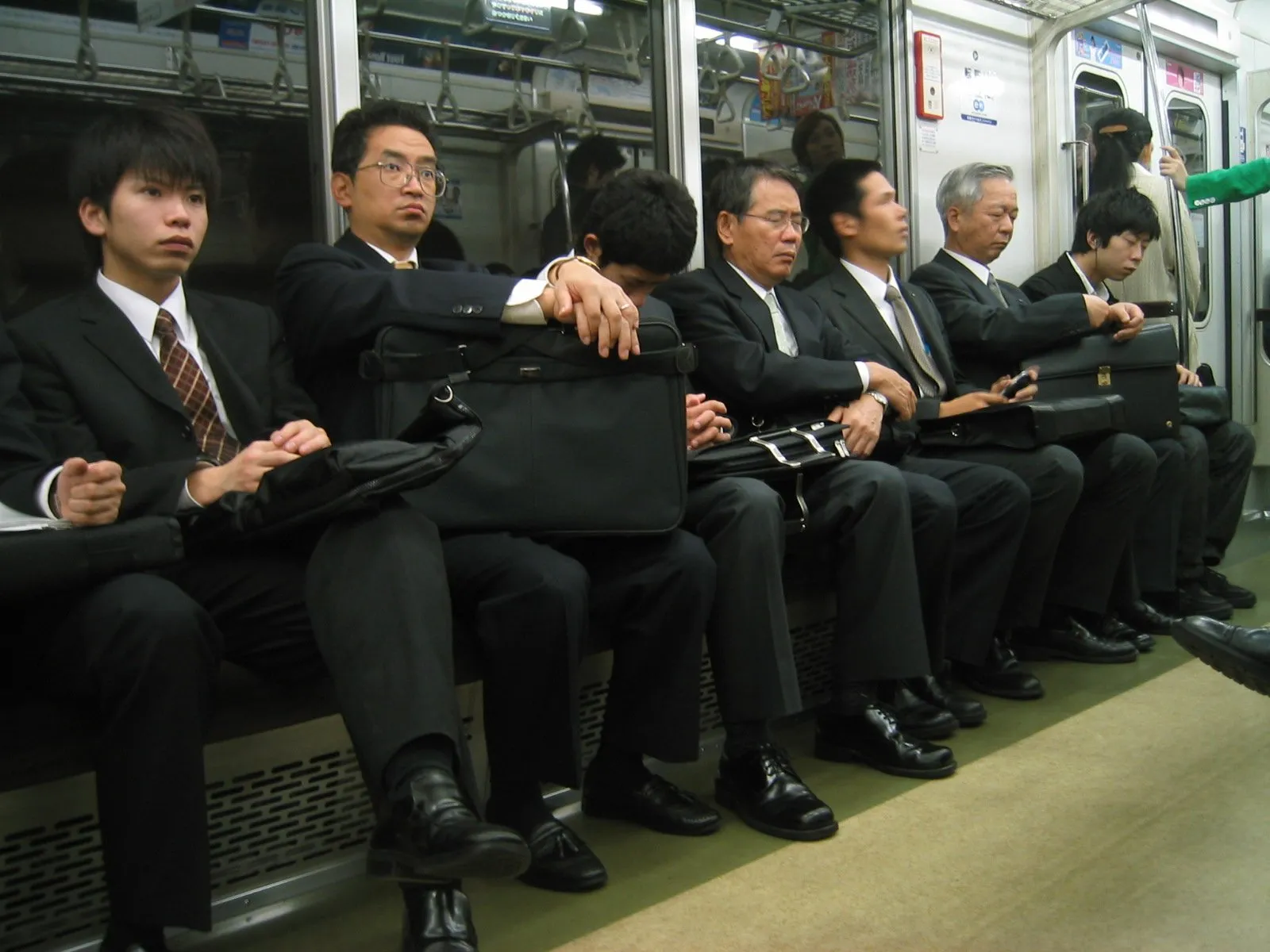 Businessmen in dark suits sitting on a train, some with handbags, others using mobile phones, all with serious expressions or sleeping, in a subway car.
